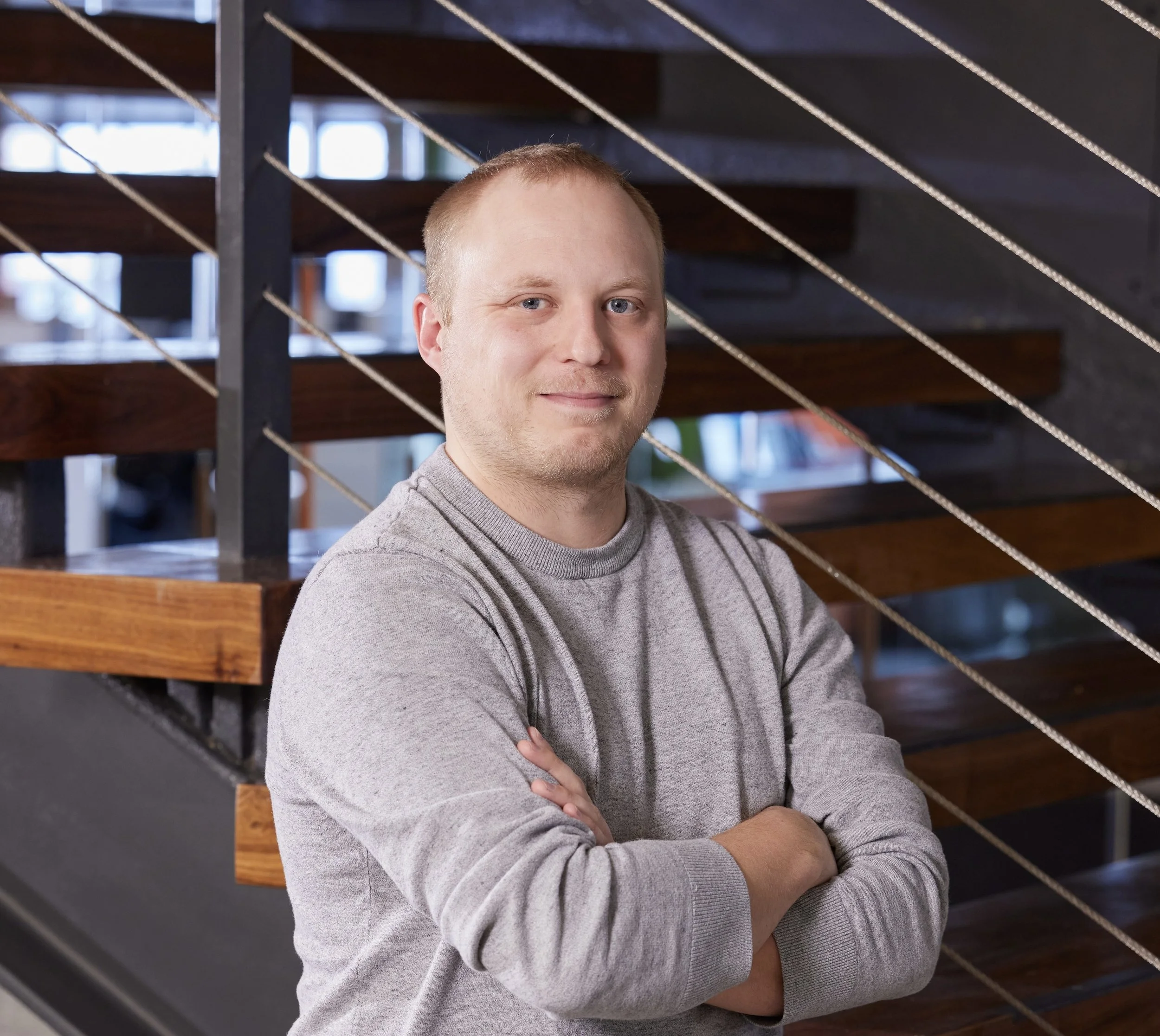 A young man with short blonde hair and a slight smile, wearing a gray sweatshirt, standing with arms crossed in front of a modern staircase with wooden steps and metal railings in an indoor setting.