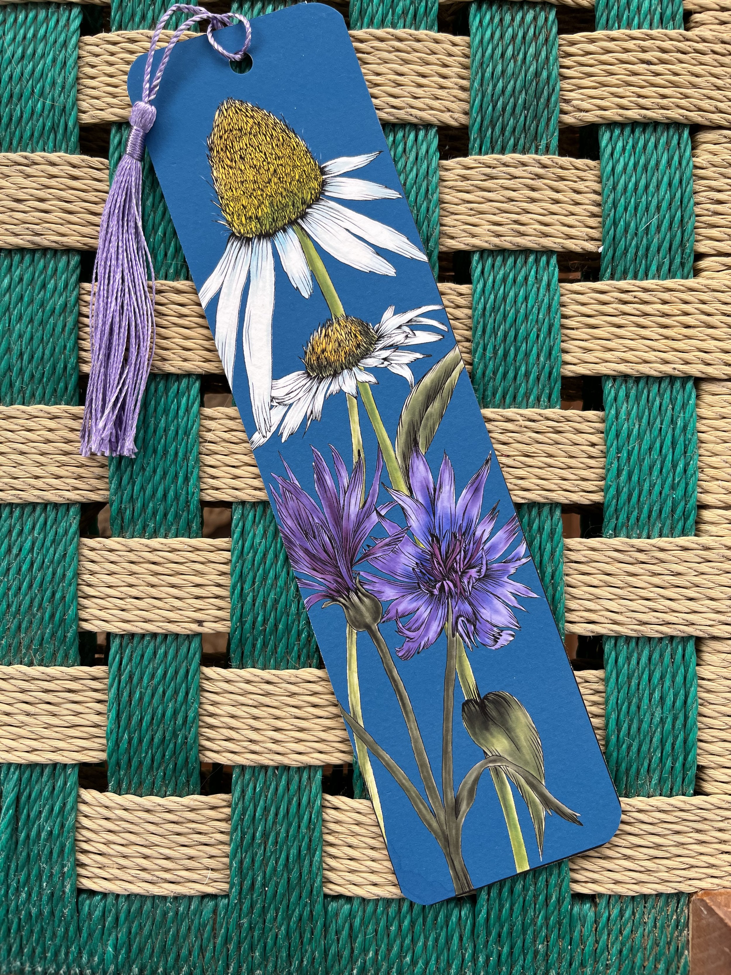 Deep blue bookmark with white daisies photographed on a wicker stool.