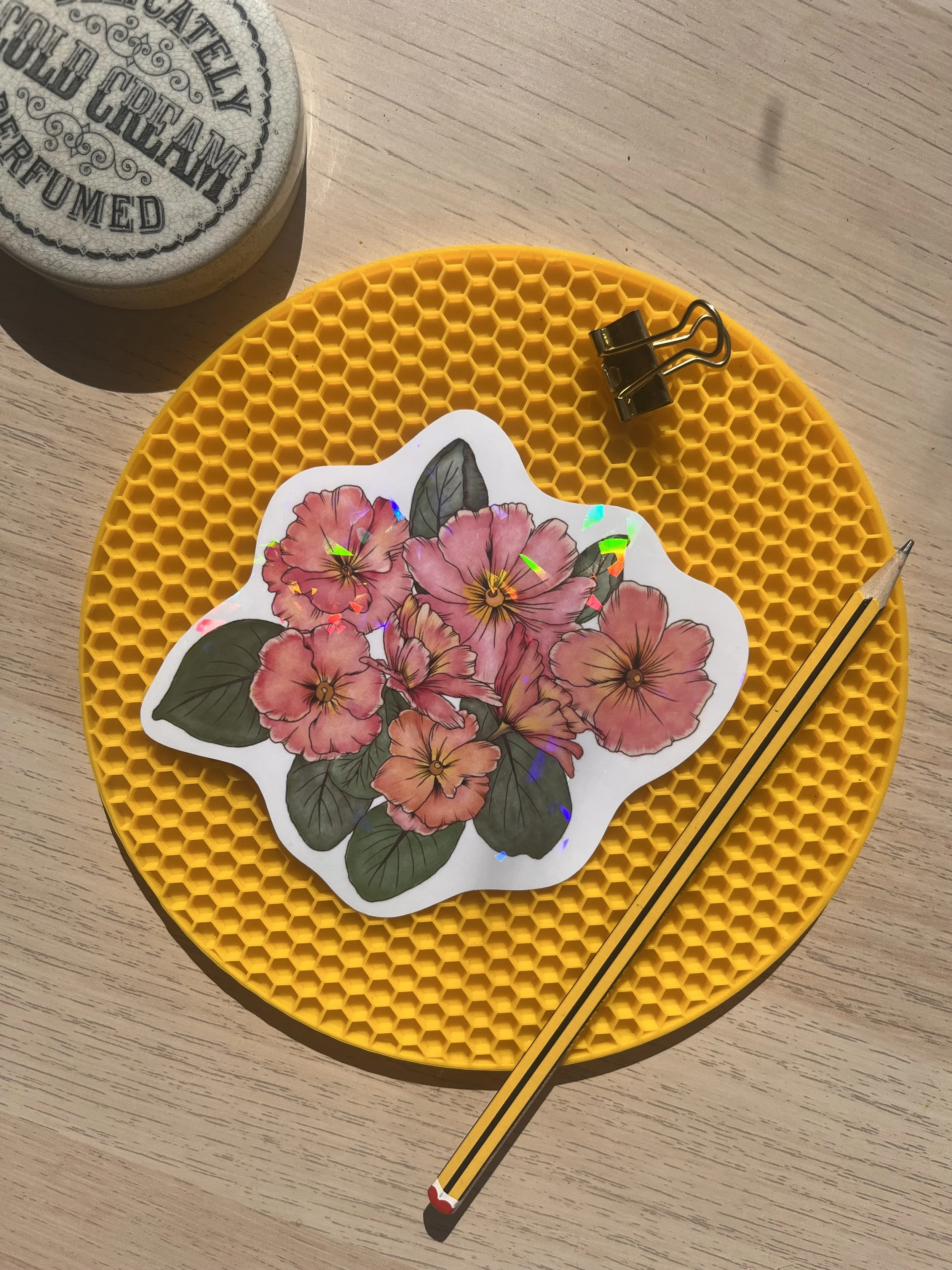 Holographic primrose sticker photographed on a yellow mat with pencil and small gold bulldog clips.