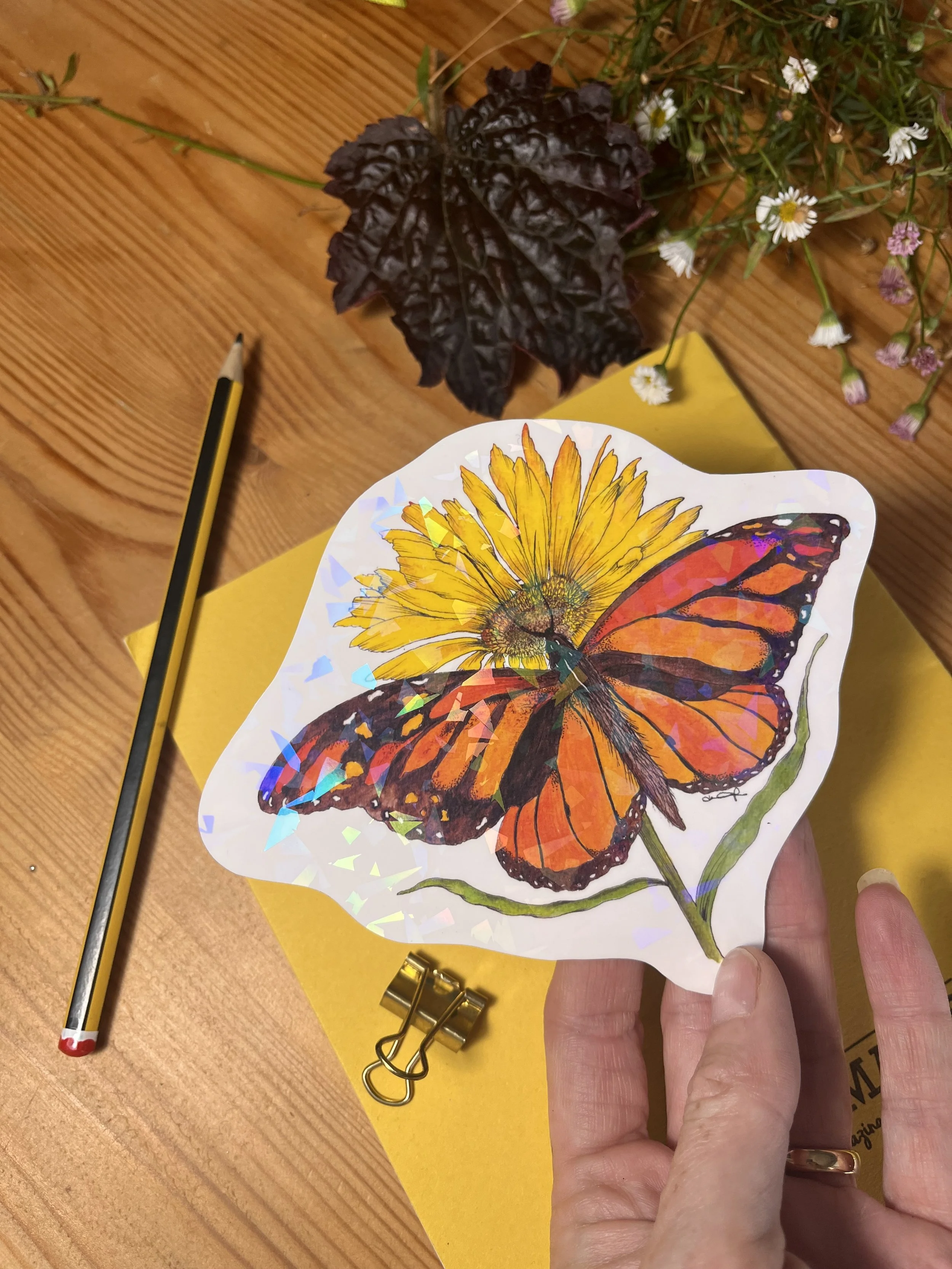 Orange butterfly holographic sticker - the sticker is held in a hand and in the background is a yellow exercise book, a pencil and a bulldog clip.