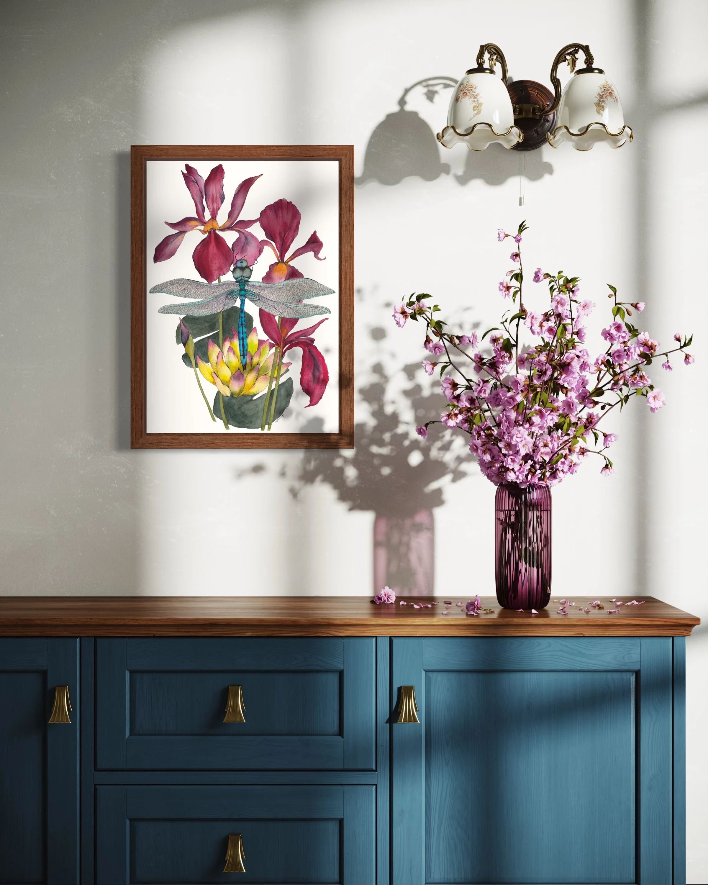 A blue sideboard with a wooden top holding a pink vase with pink cherry blossoms, a framed artwork of a dragonfly and flowers on the wall, and a brass and glass light fixture above.