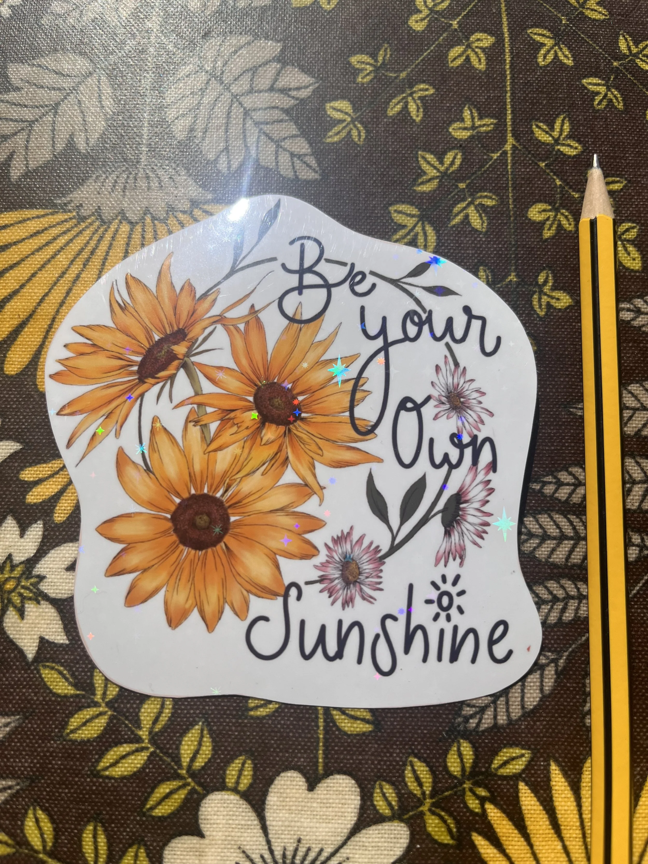 Holographic yellow sunflower sticker - the sticker is photographed front facing against a floral tray with a pencil to one side.