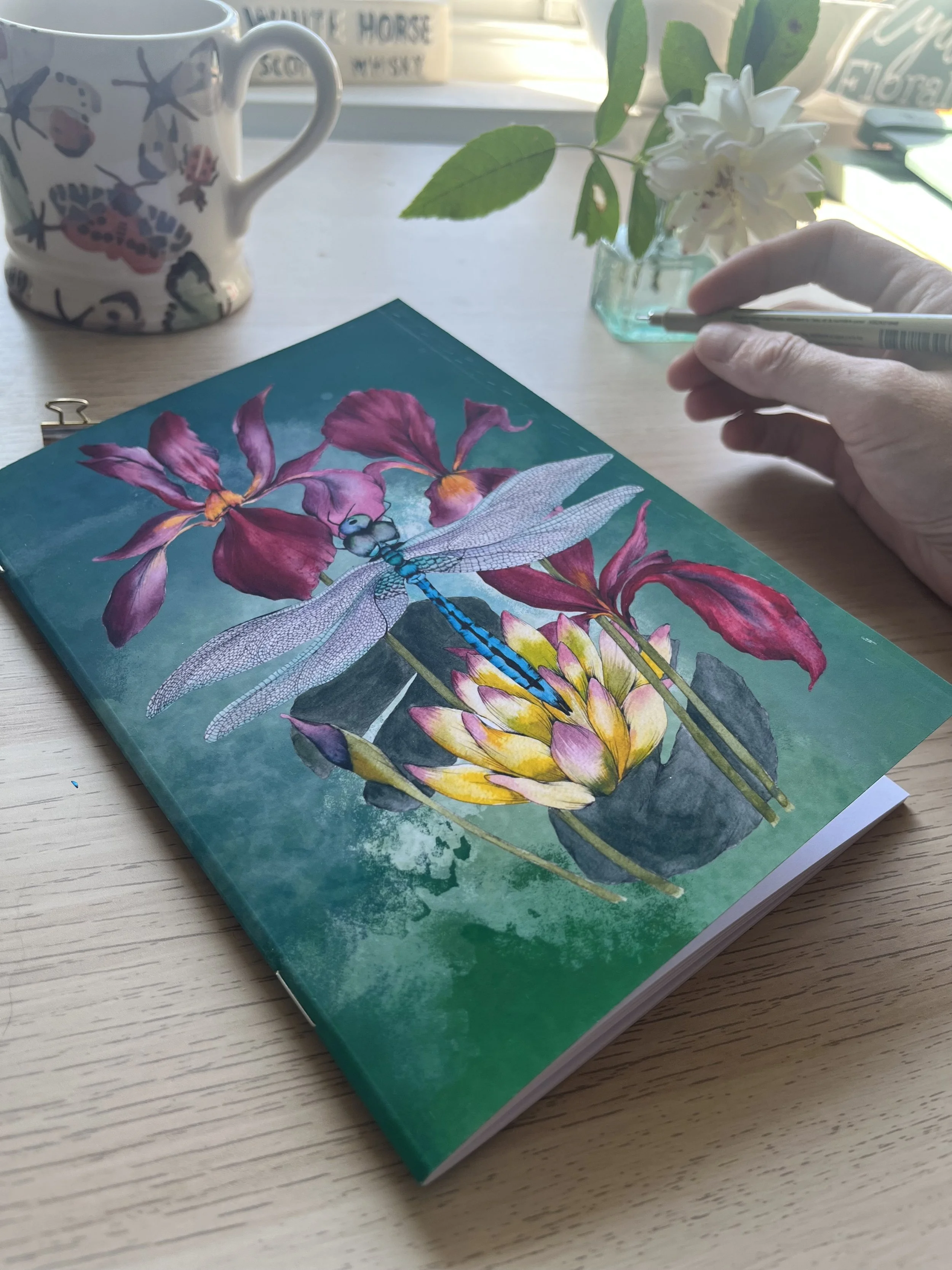 Deep green and blue Notebook with dragonfly on the cover. Photographed with a hand holding a pen and flowers