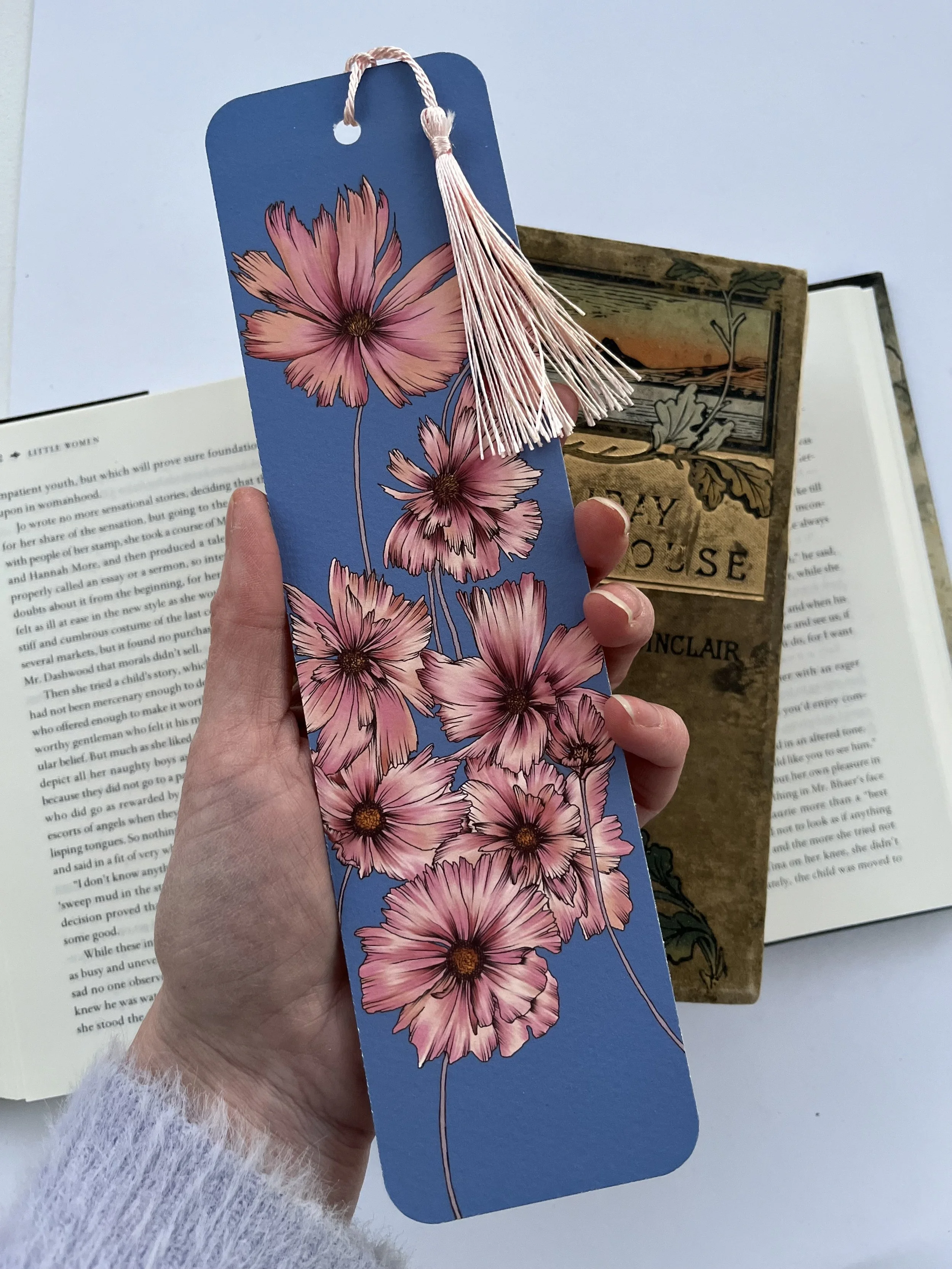 A blue tassel bookmark with pink flowers photographed held in a hand with some vintage books in the background.