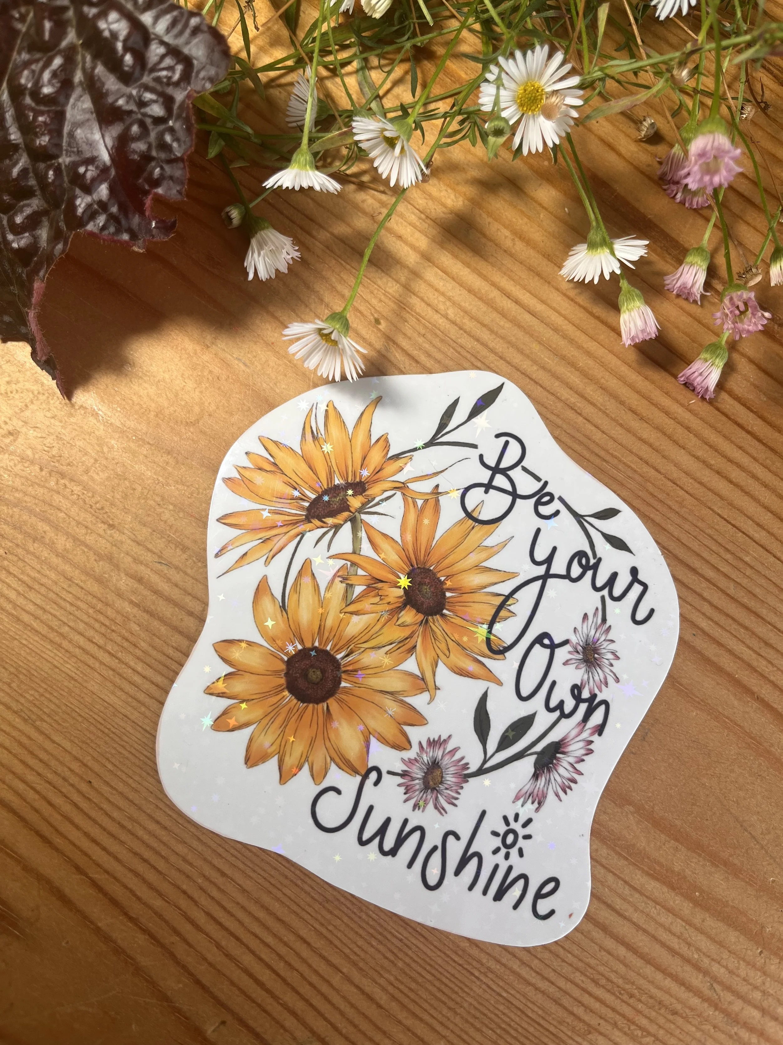 Sunflower holographic Daisy sticker photographed on a wooden table surrounded by flowers
