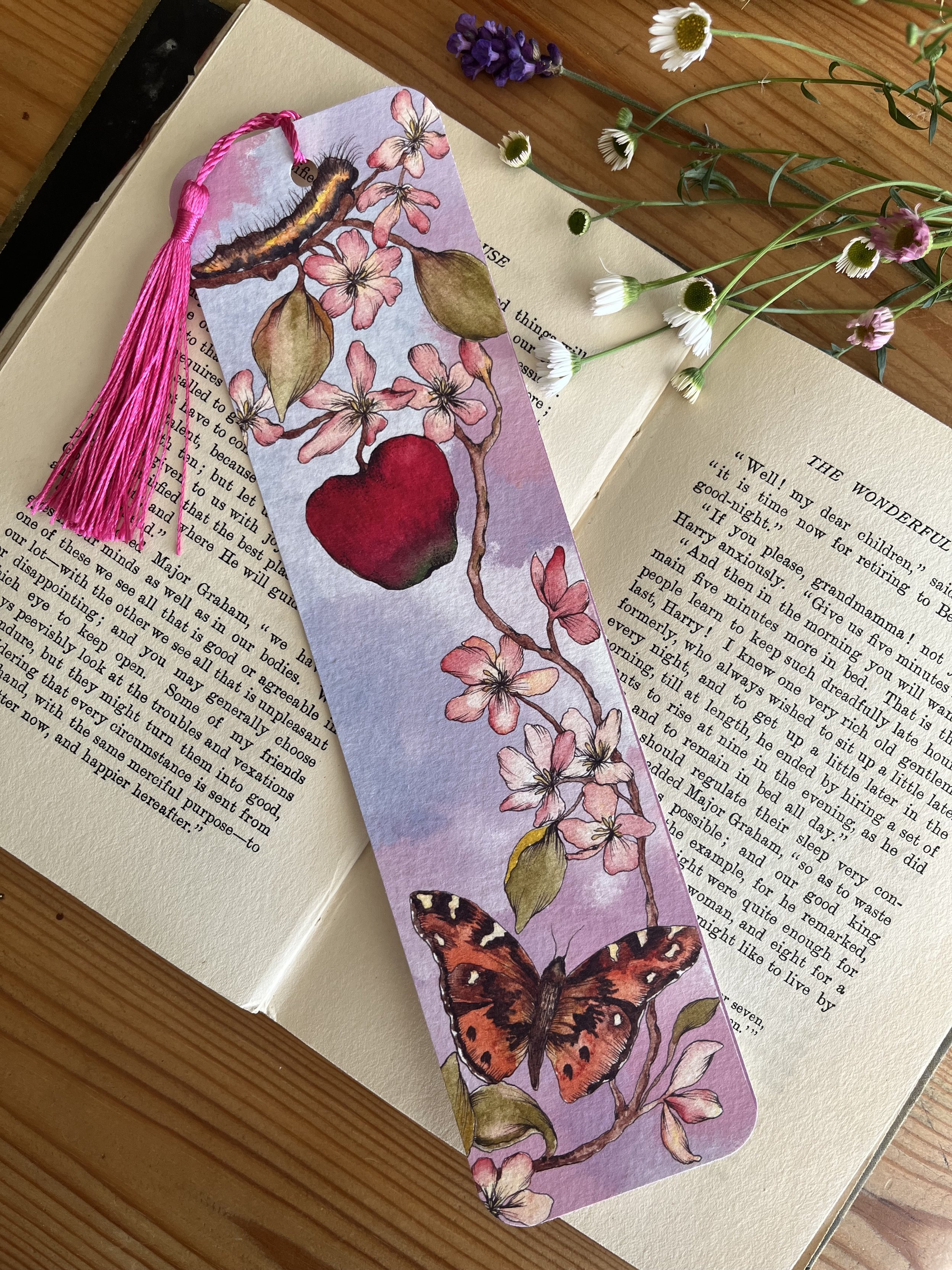 A pale pink tassel bookmark with flowers photographed on an open book with flowers in the background.