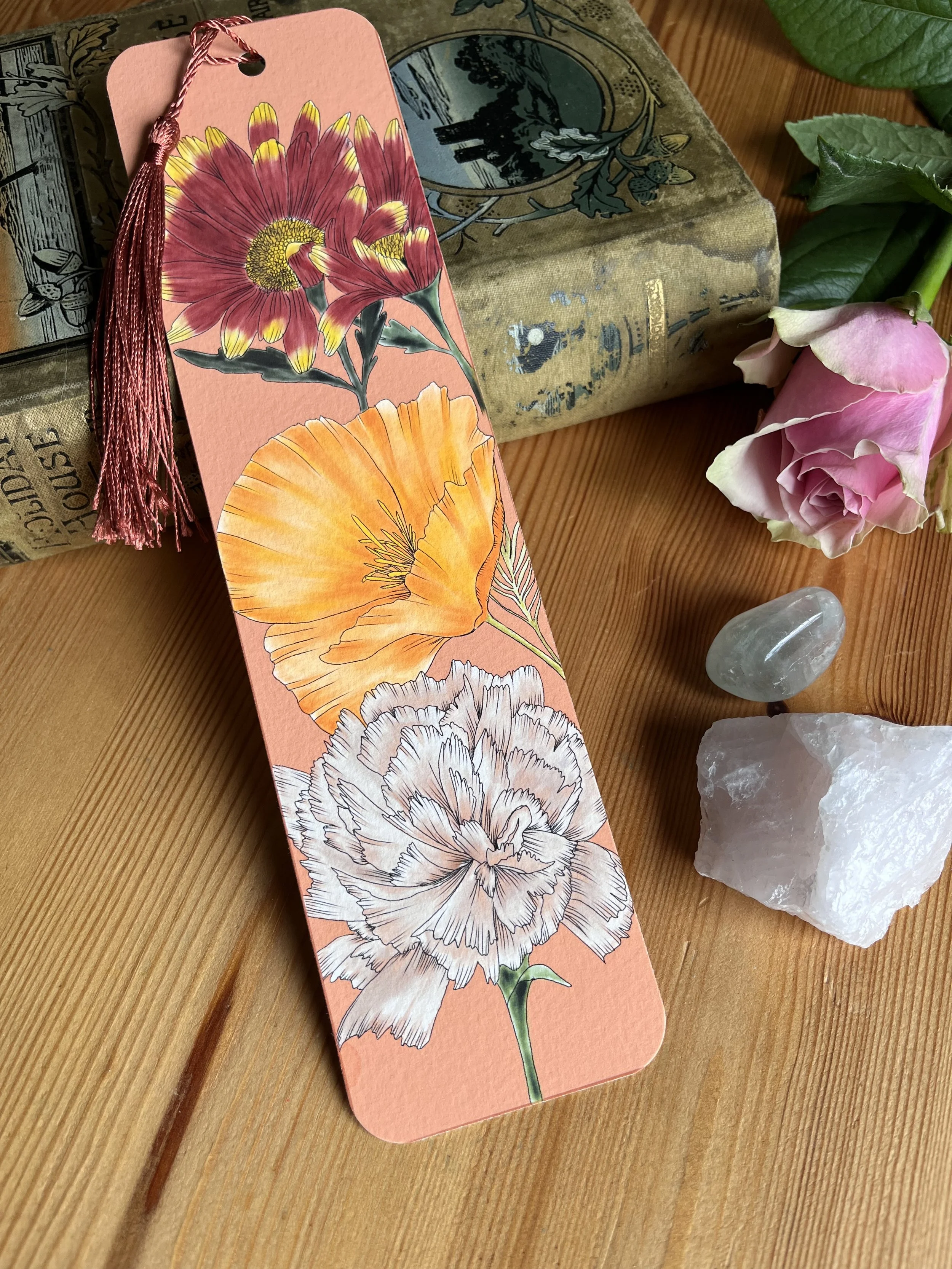 An orange tassel bookmark with carnation flower photographed on a wooden table with a rose and crystals