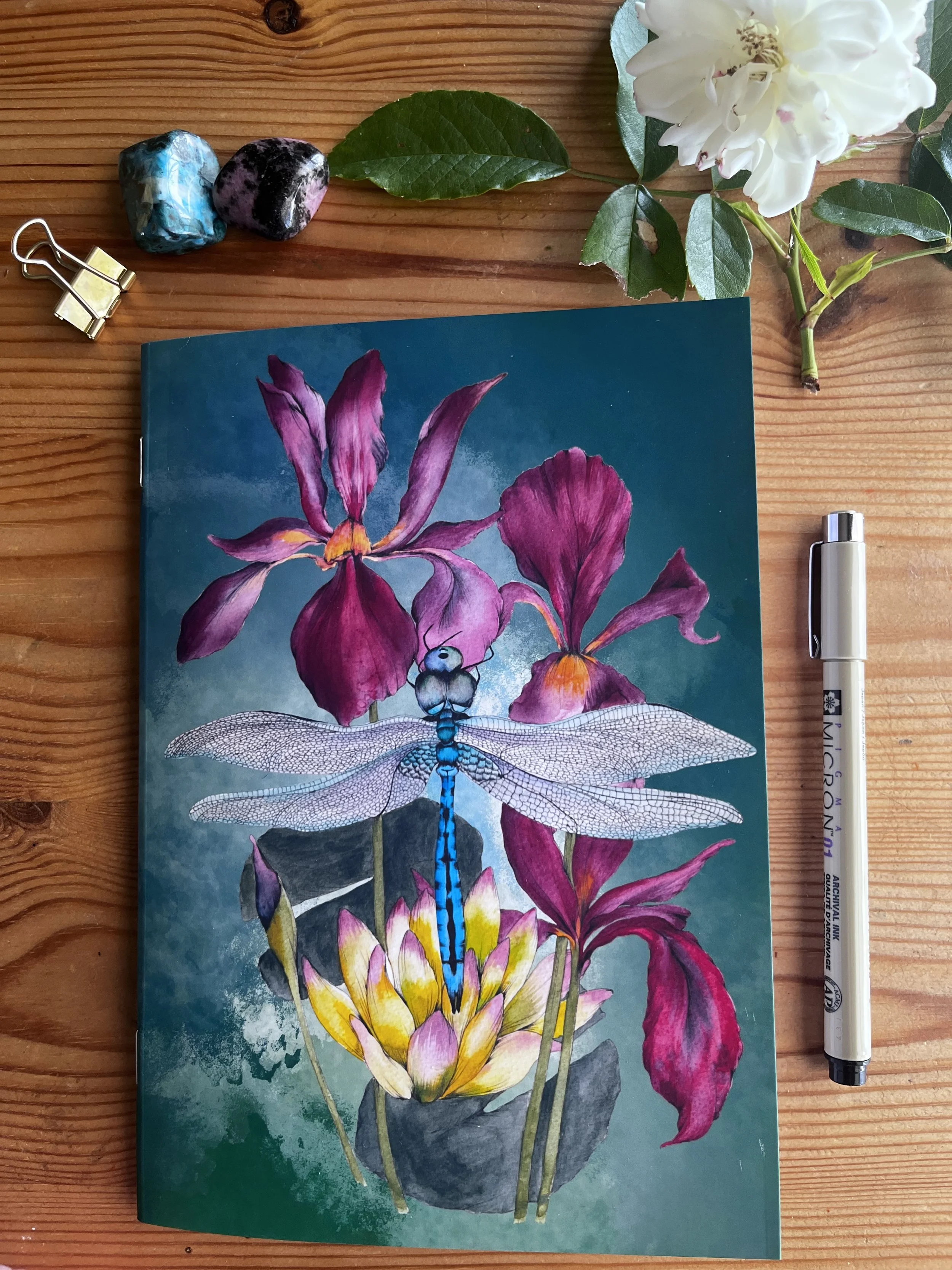 Green dragonfly notebook is pictured on a wooden background with crystals and flowers