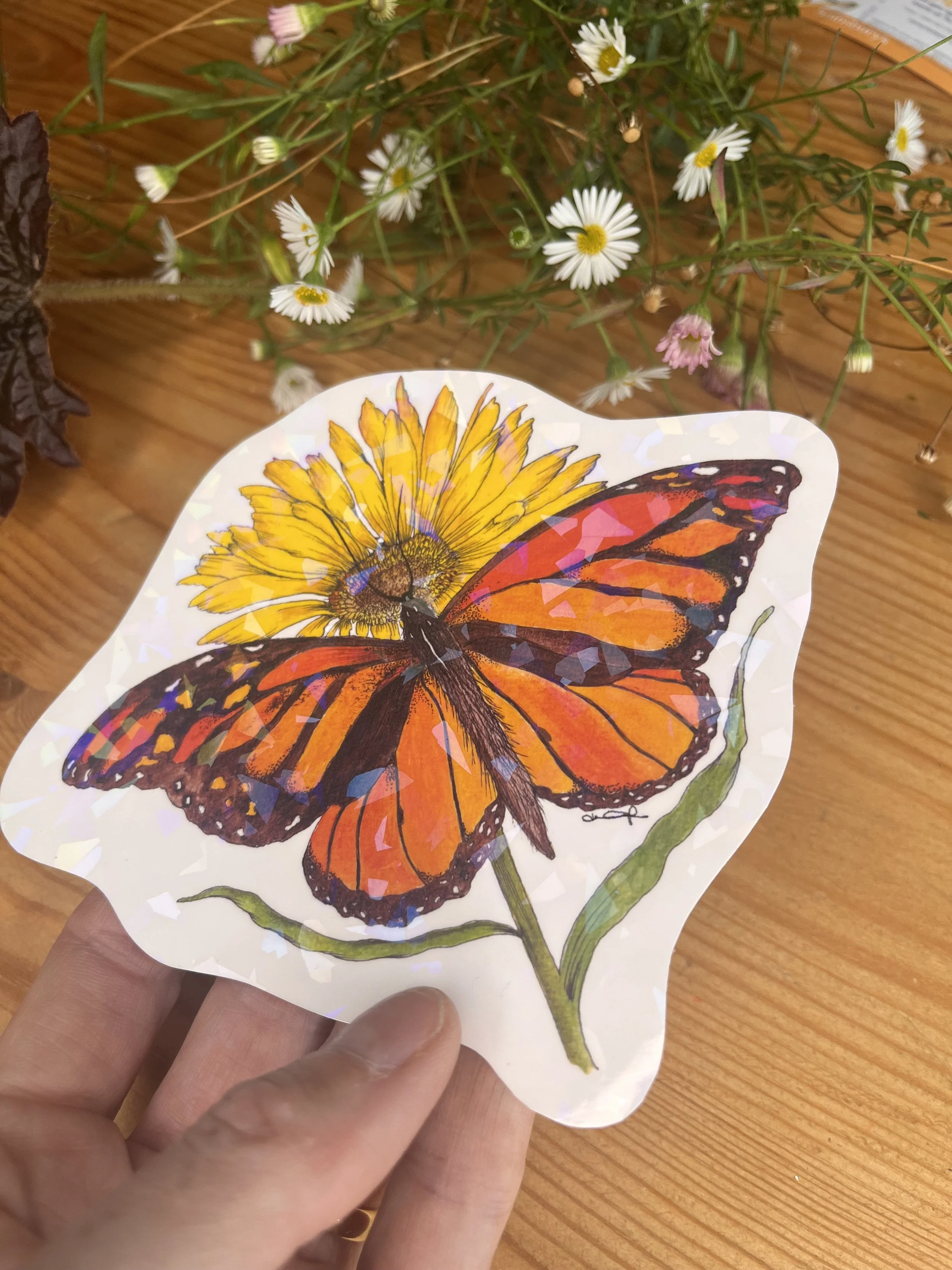 Orange butterfly sticker held in a hand on a wooden table with daisy flowers surrounding it.