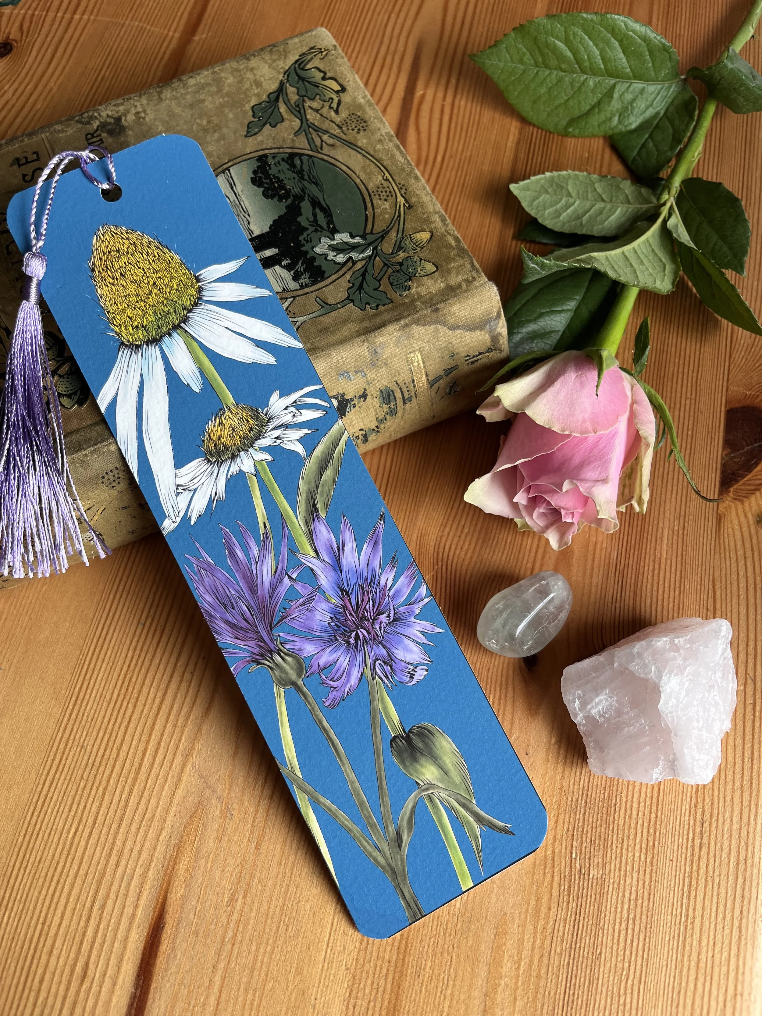 Deep blue bookmark with a large white daisy. Photographed against a wooden backdrop with a rose and crystals
