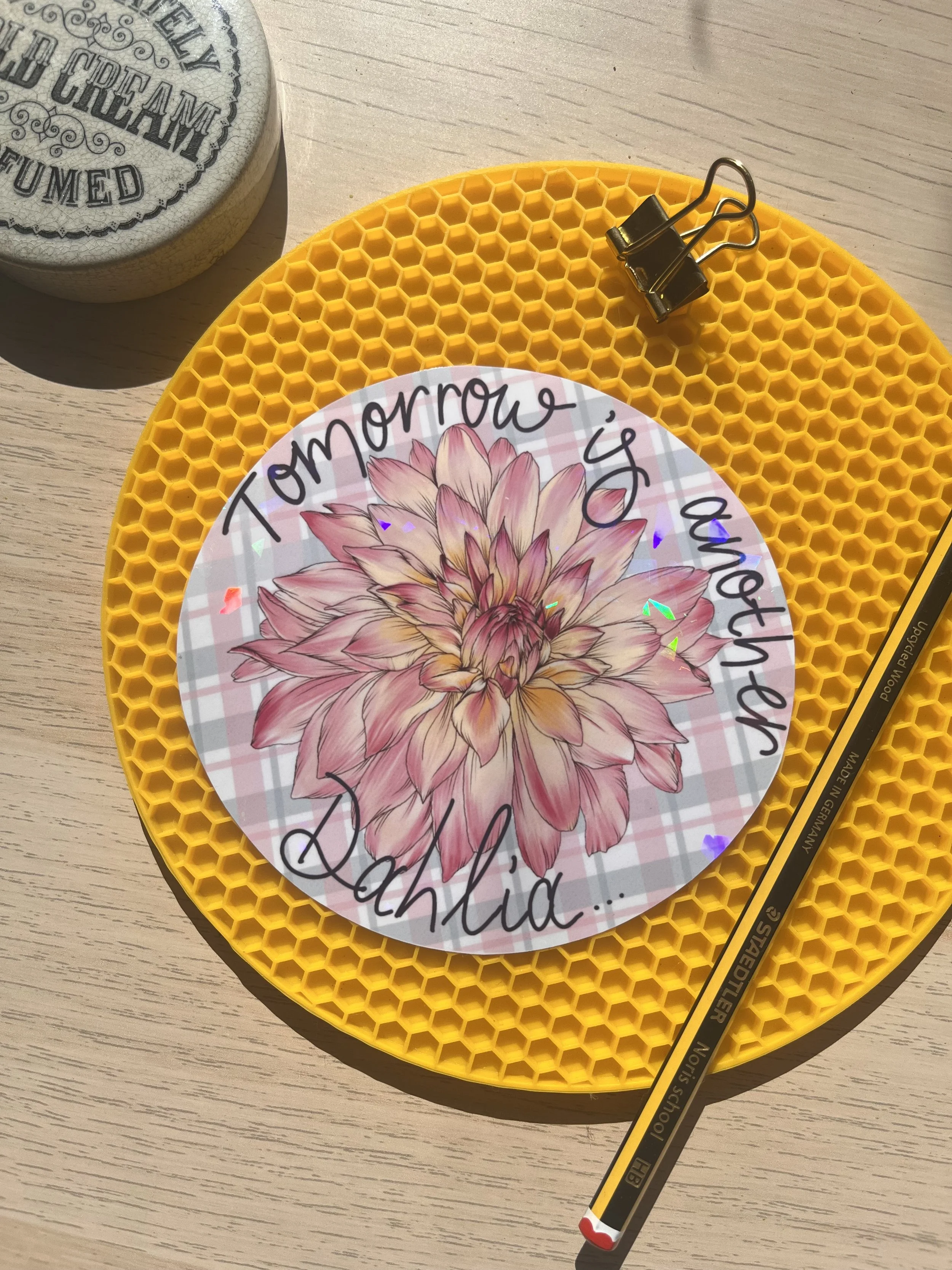 Holographic circular sticker with dahlia photographed against a circular yellow mat with a hub pencil and bull dog clips in the background.