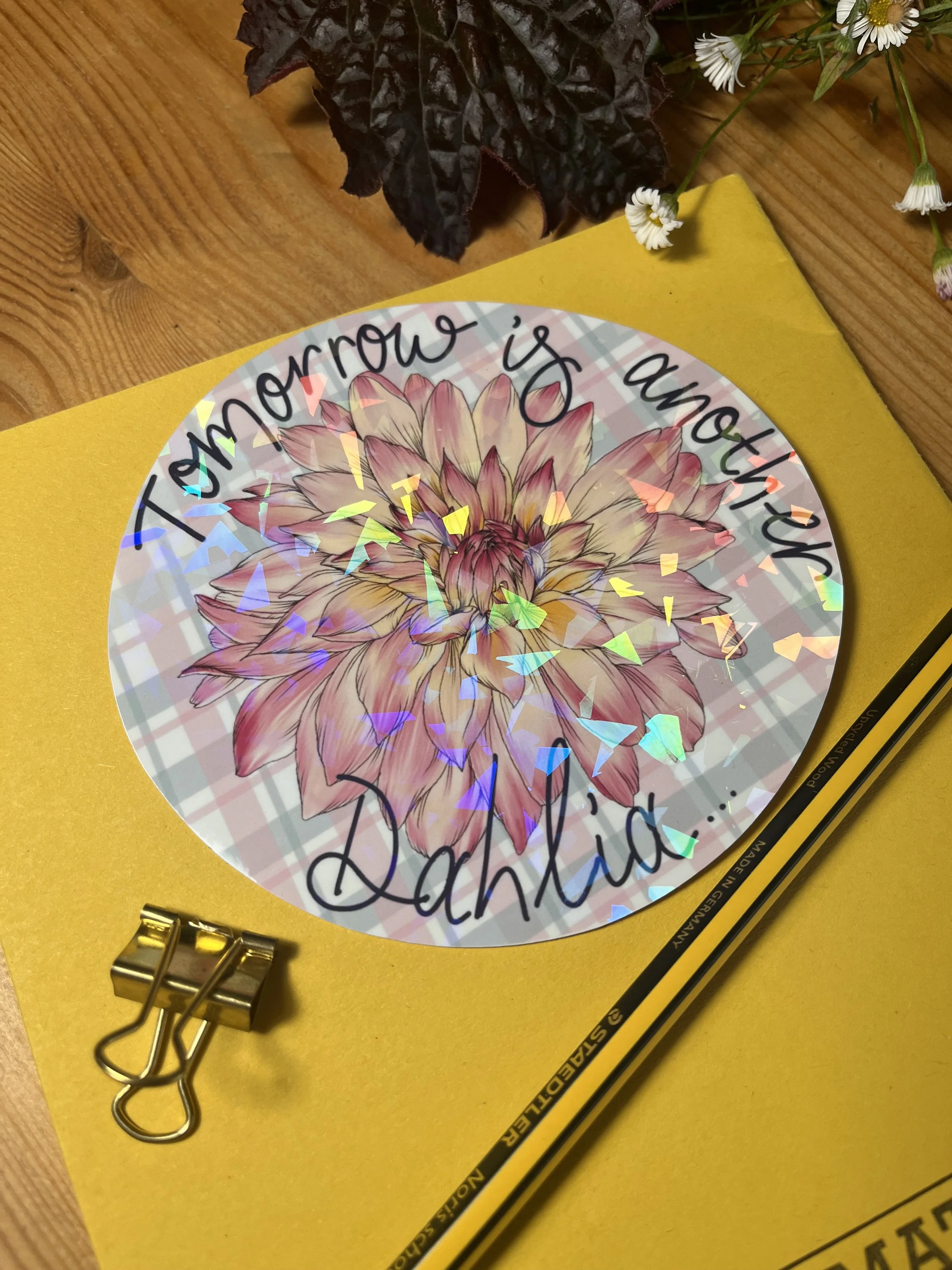 Holographic circular pastel sticker with dahlia.the sticker is photographed on a yellow exercise book and bulldog clips are shown next to the sticker.