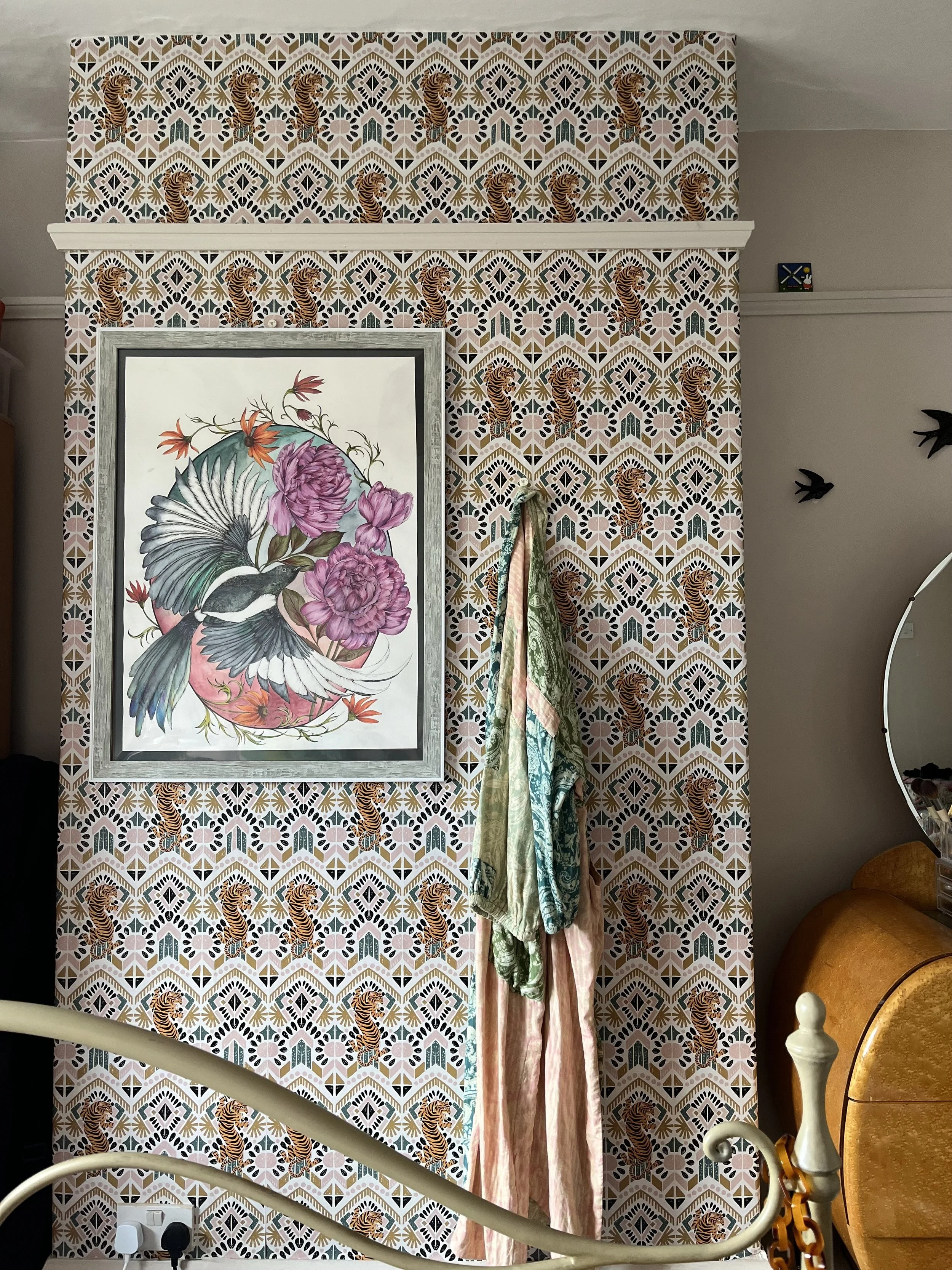 A room wall with patterned wallpaper featuring tiger and geometric designs, a framed floral and bird artwork, and hanging fabrics. A mirror and part of a bed frame are also visible.