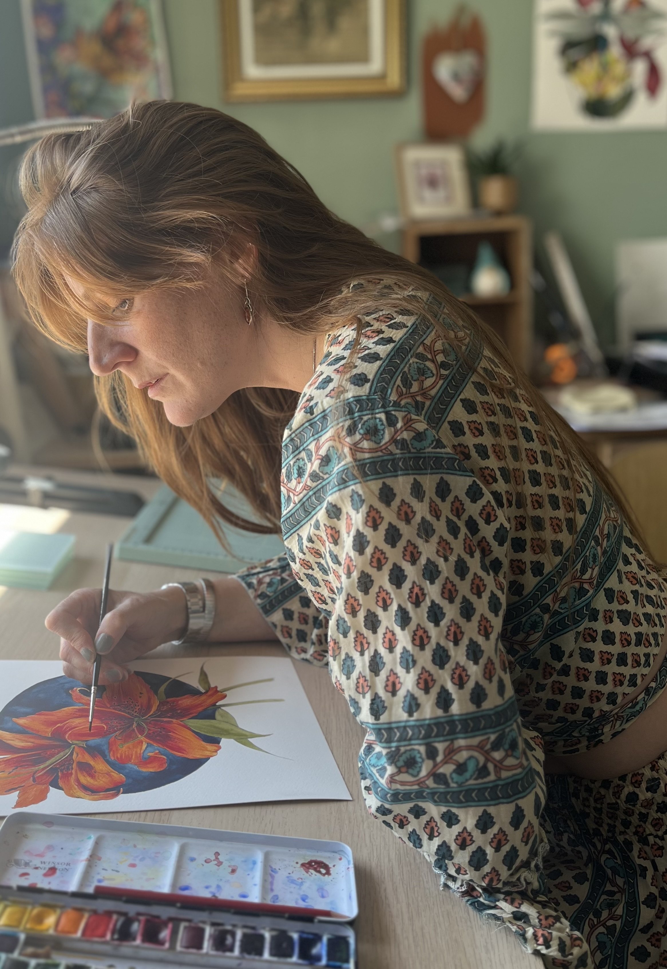 Woman painting a vibrant orange and red flower on a white paper, using watercolor paints in an art studio.