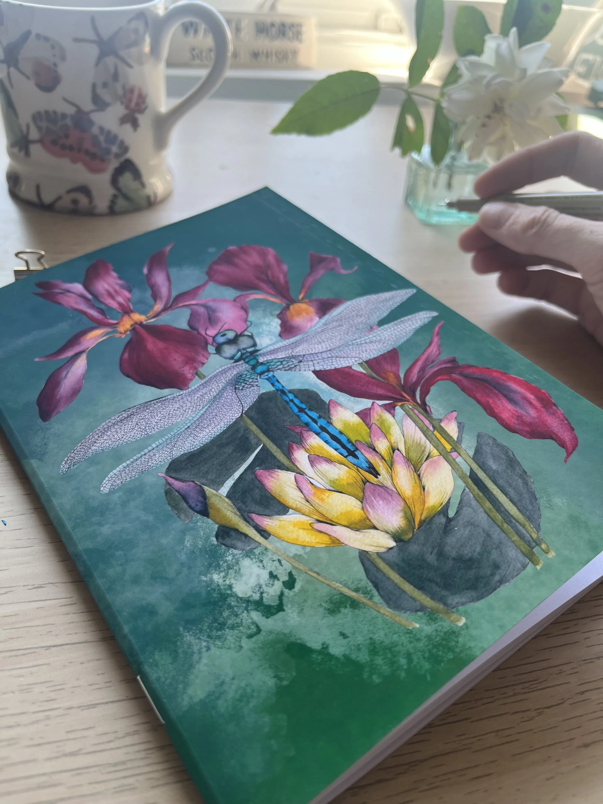 Deep green and emerald dragonfly notebook photographed on a table with a hand holding a pen and flowers in the background