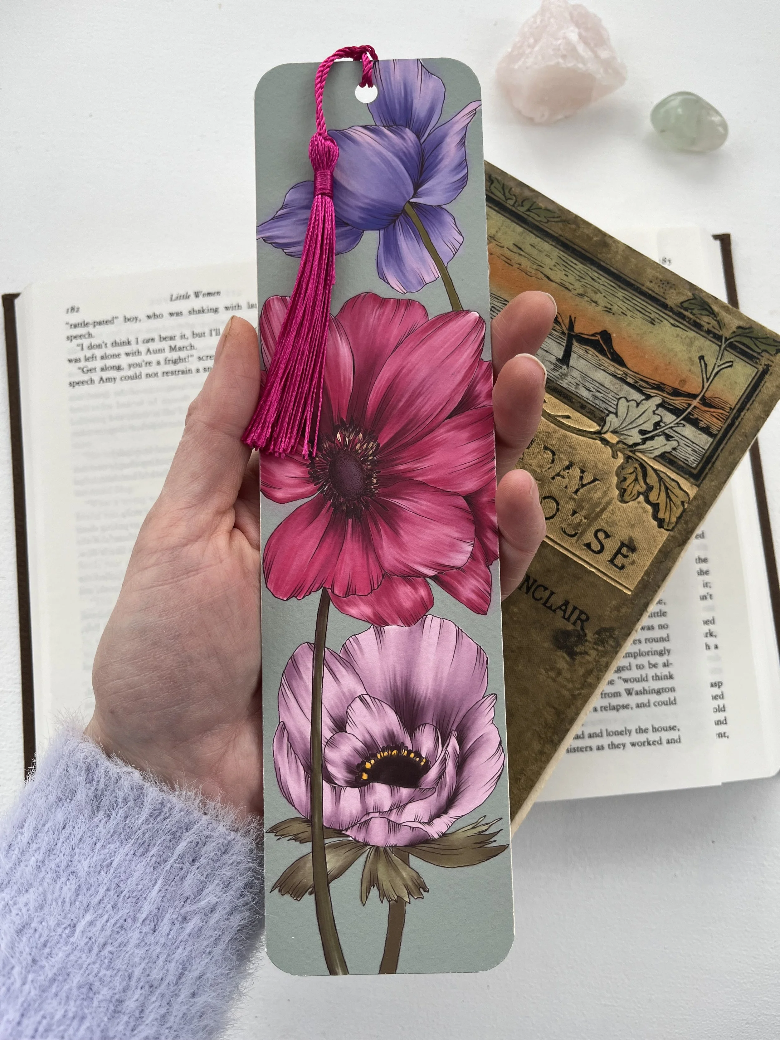 A hand holding a bookmark with colorful flowers, including purple, pink, and lavender, with a pink tassel. In the background are books and decorative stones.