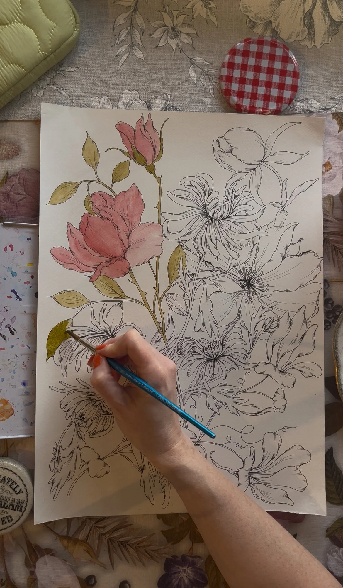 A person is painting a floral illustration on paper with watercolors, highlighting pink flowers and green leaves, surrounded by art supplies and floral-patterned fabric.