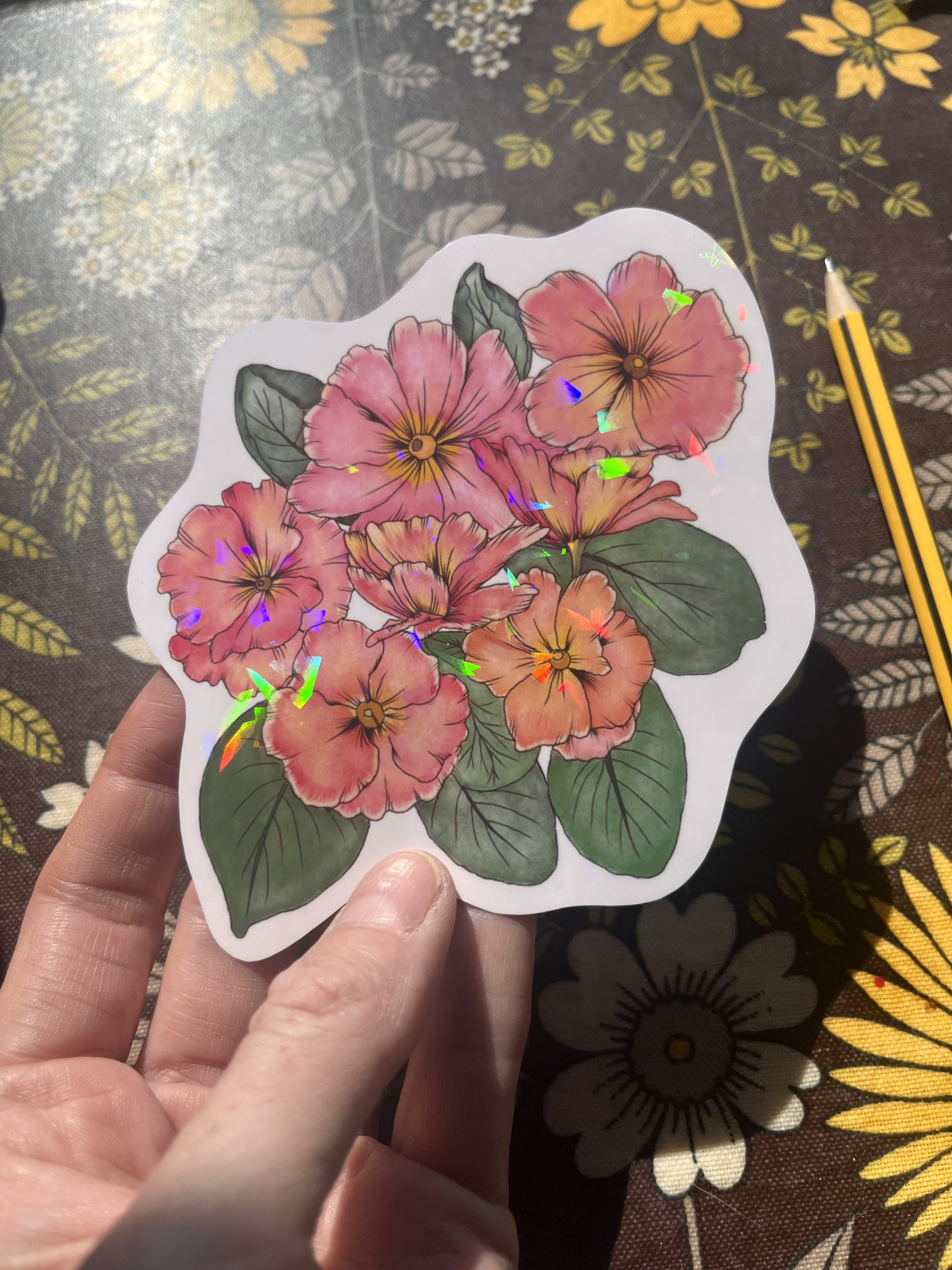 Holographic primrose vinyl sticker - the sticker is held in a hand showing the reflective nature of the sticker. The background is a brown floral tray.