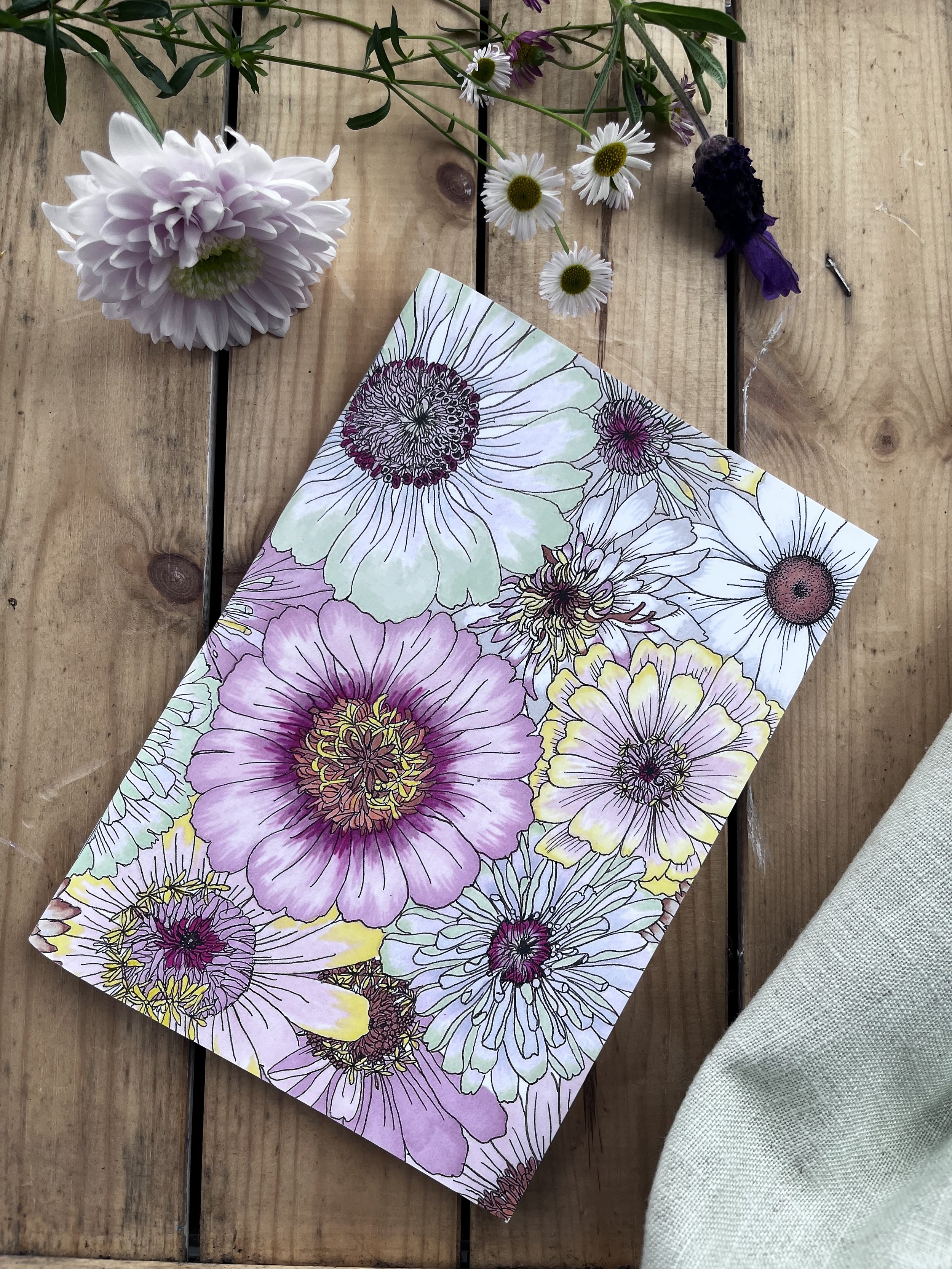 Pale pastel notebook photographed on a slant on wooden boards. There are flowers in the background.