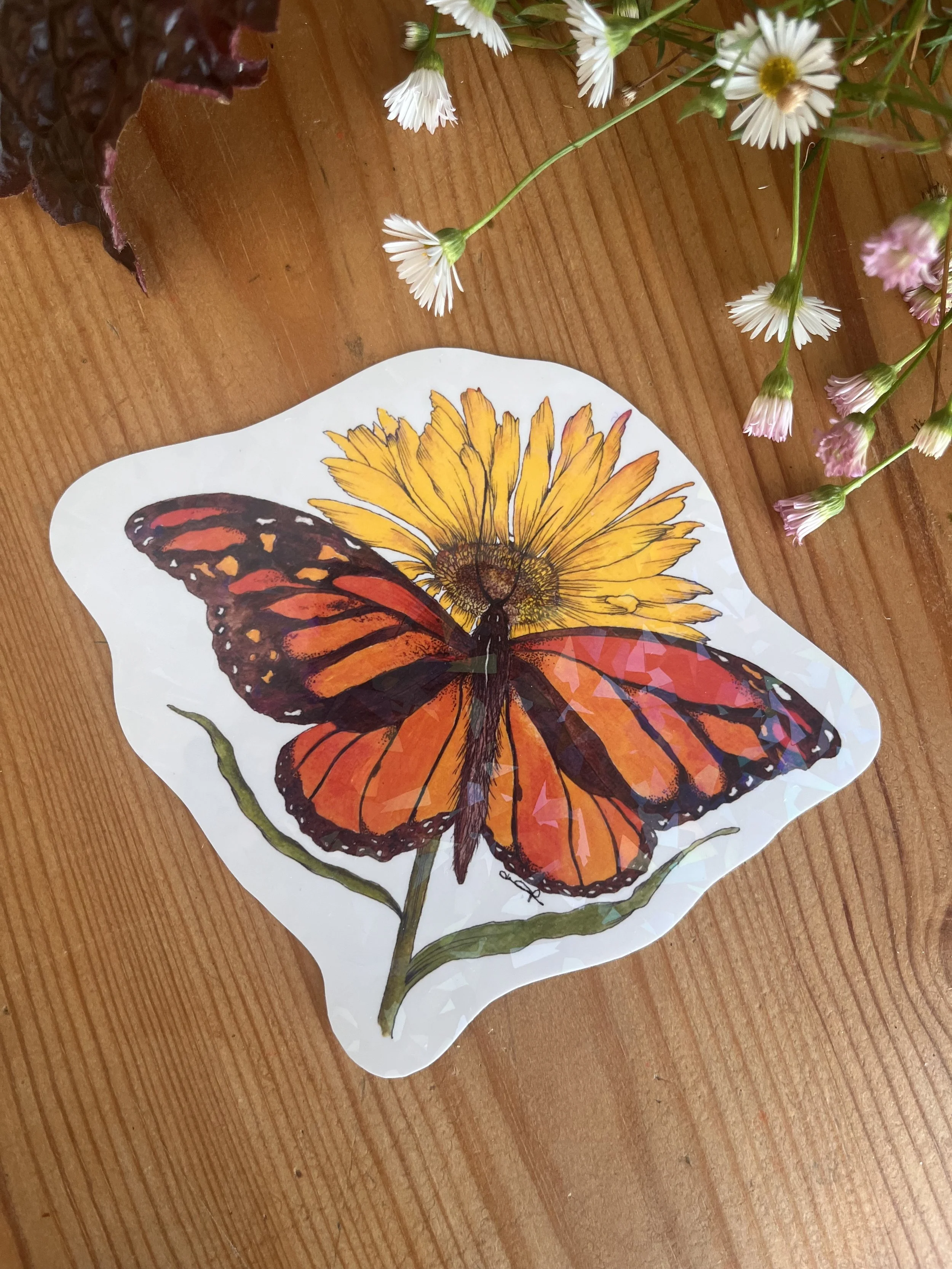 Orange monarch butterfly sticker on a wooden table with daisy flowers surrounding it.
