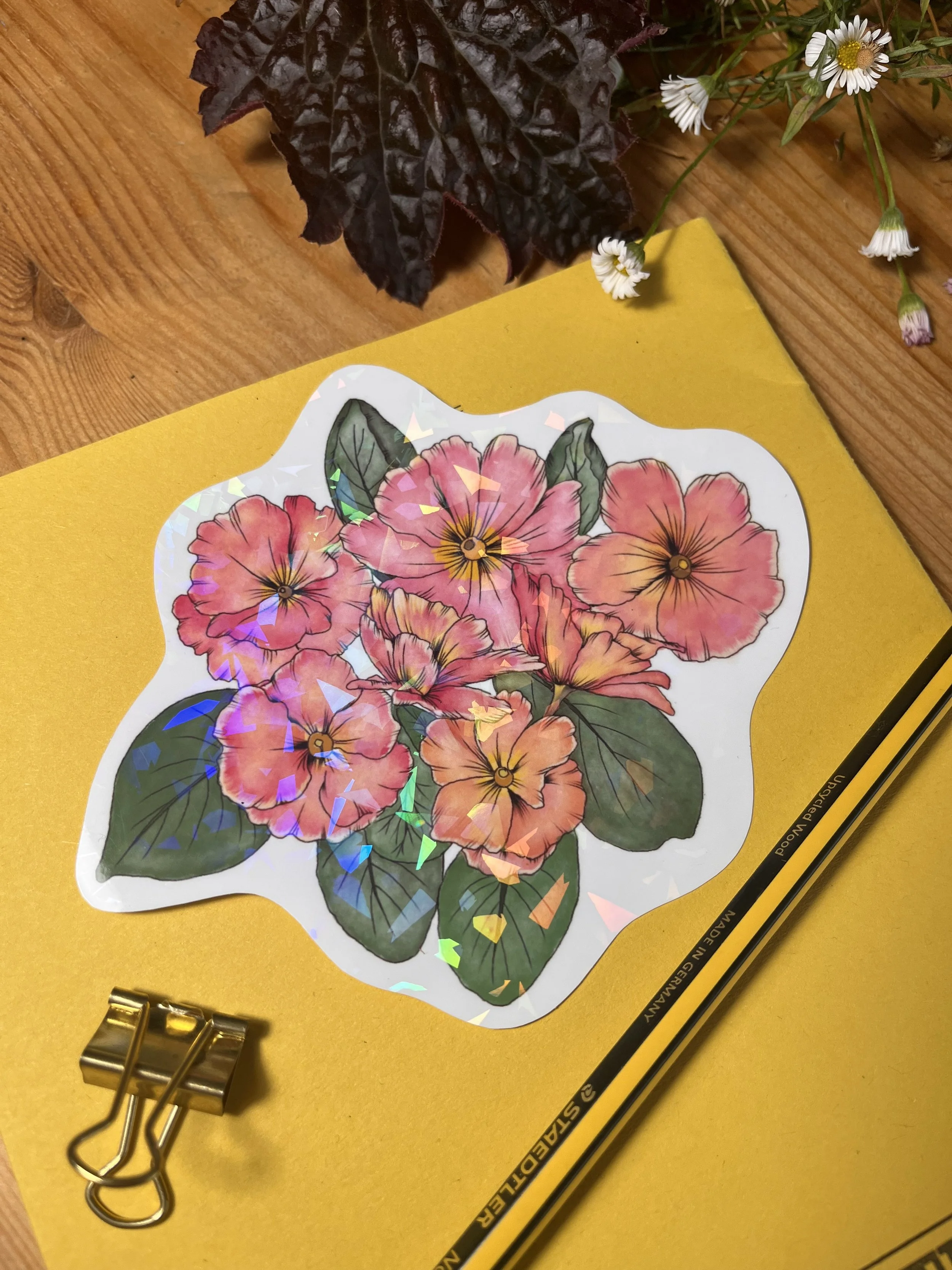 Holographic primrose sticker photographed on a yellow exercise book with a pencil in the background.