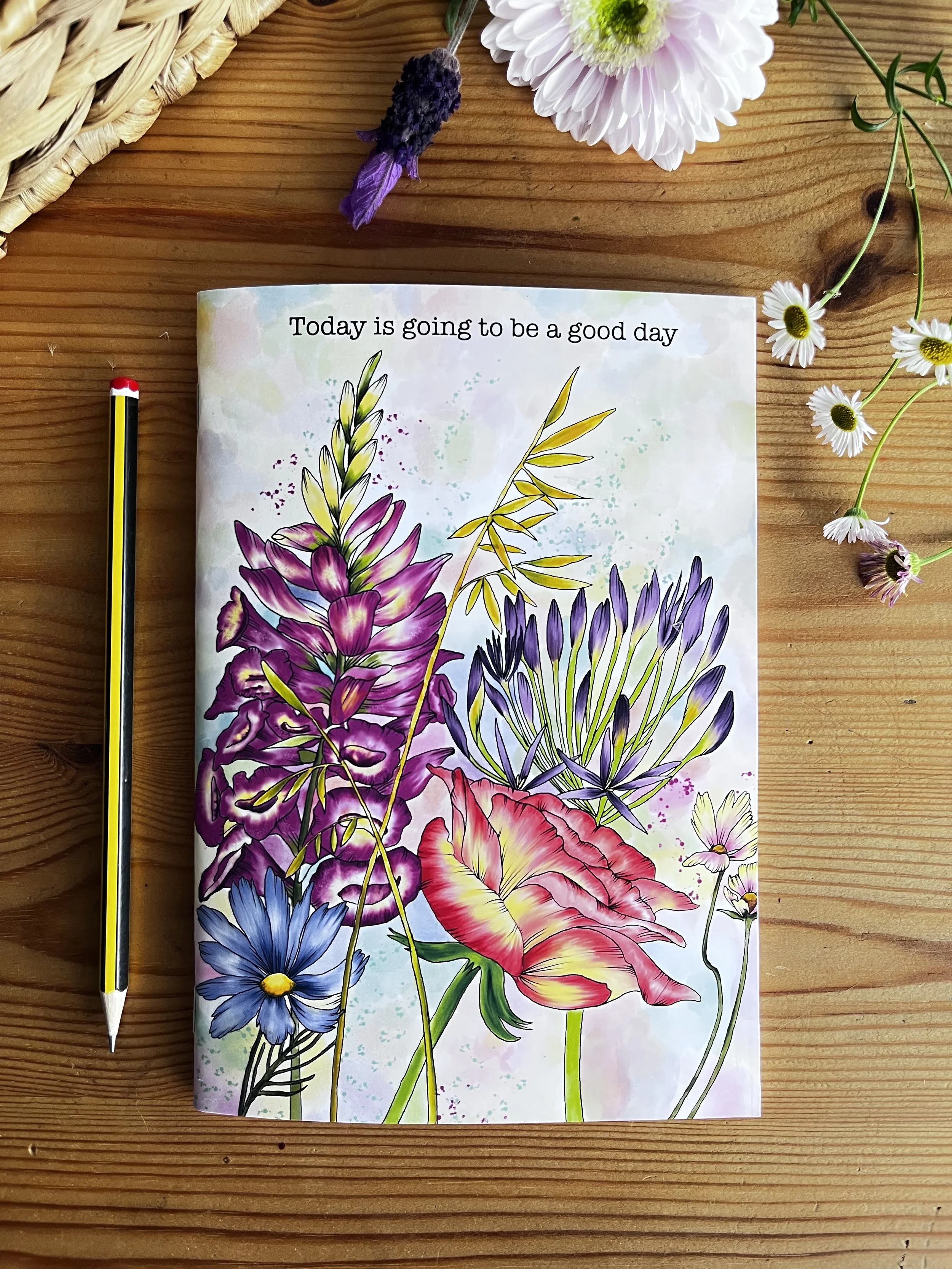 Wildflower notebook with pink flowers, photographed on a wooden table with a hub pencil and a wicker hat in the background
