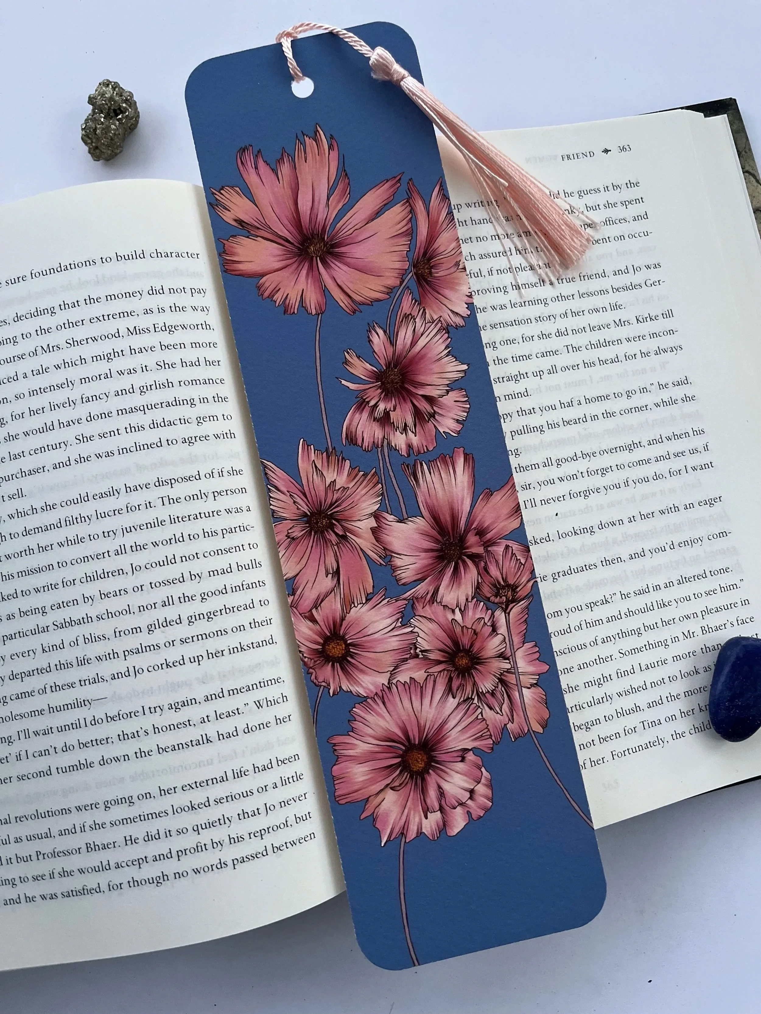 Blue bookmark with tassel and pink flower design. Photographed on an open books against.