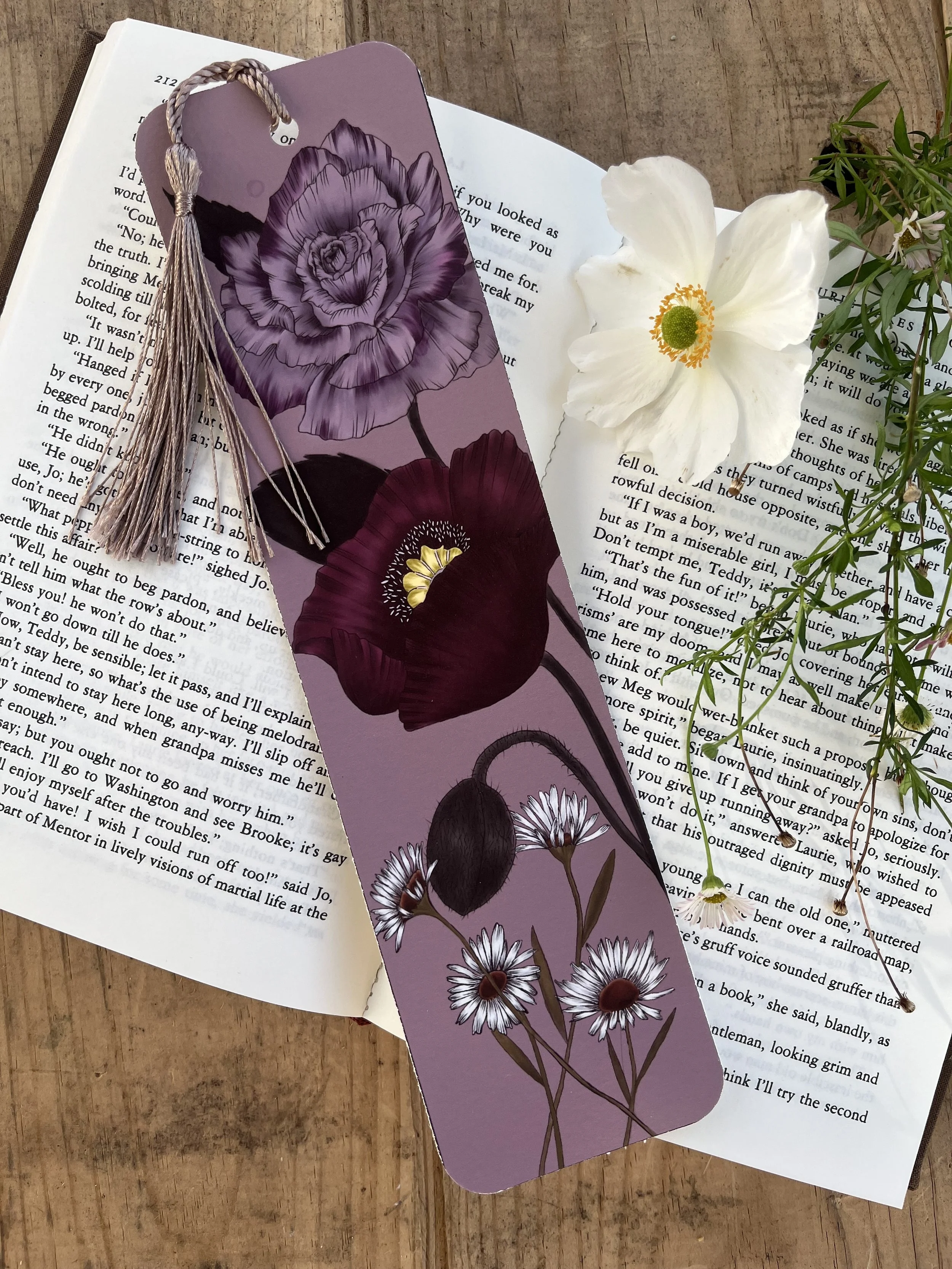 Open book with a floral bookmark, white flower, and greenery on a wooden surface.