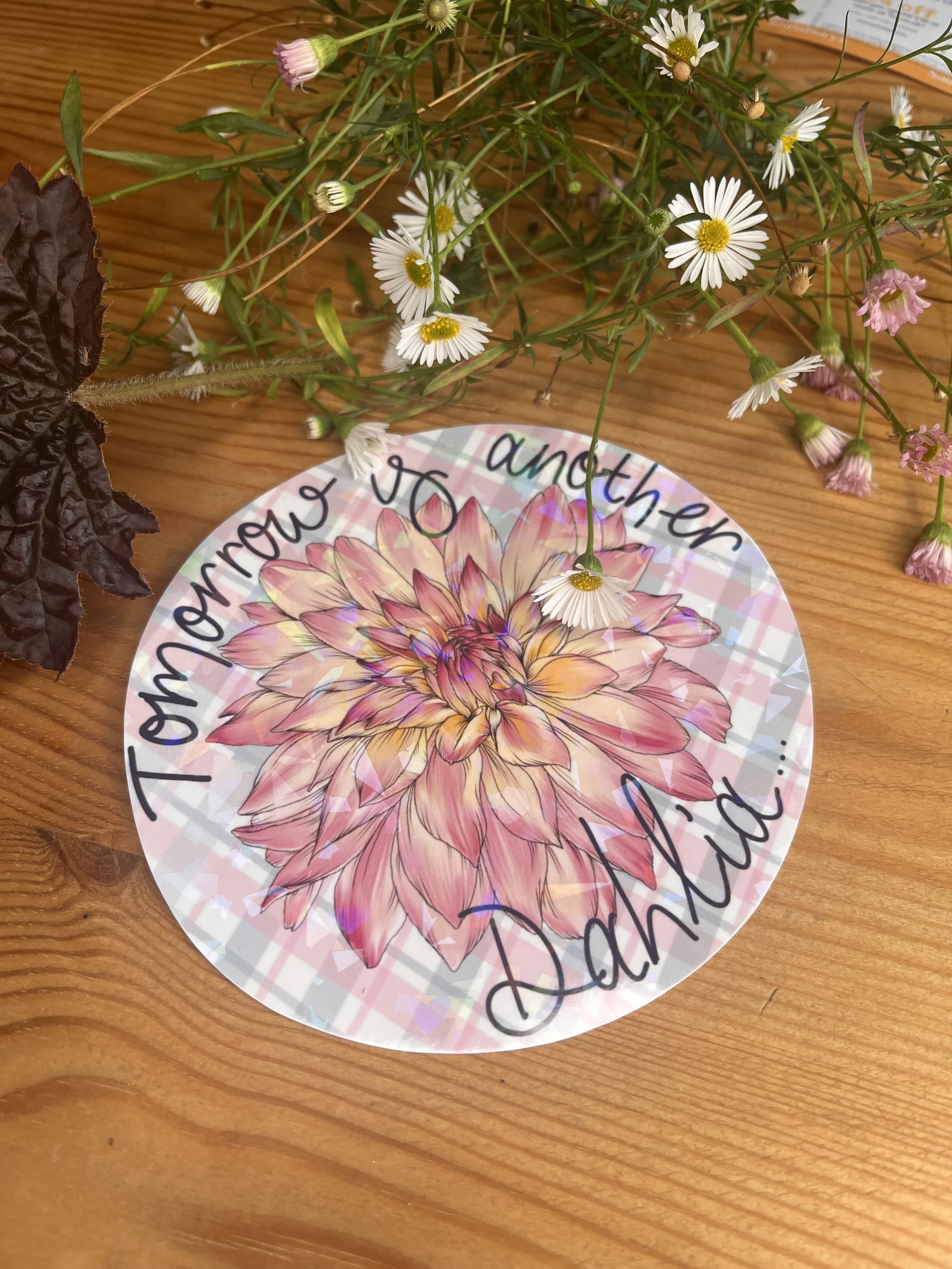 Holographic dahlia sticker. The sticker is circular photographed from the front view against a wooden table and there are flowers in the background.