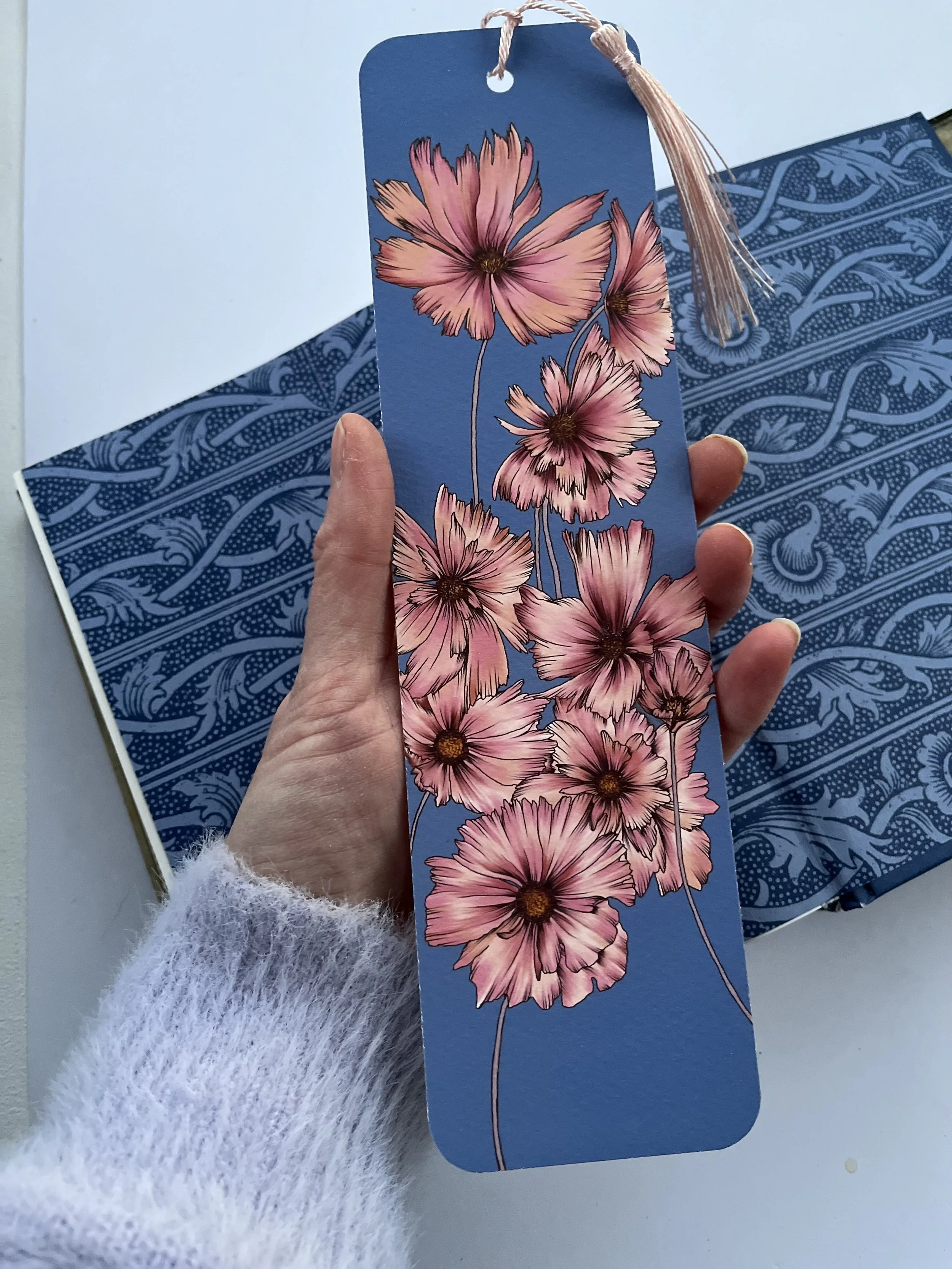 Blue bookmark with pink flowers and tassel, photographed held in a hand with a blue book behind.