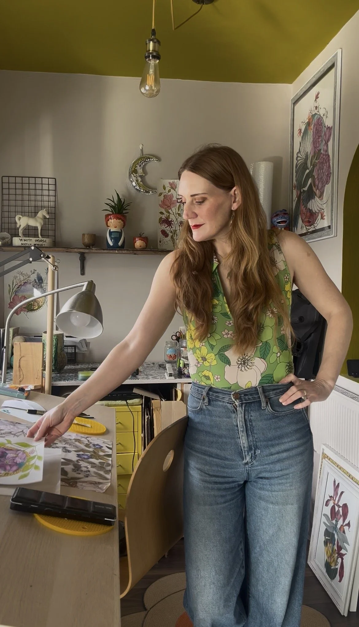 A woman with long red hair and floral green top working at a study table in a colorful, artful room.