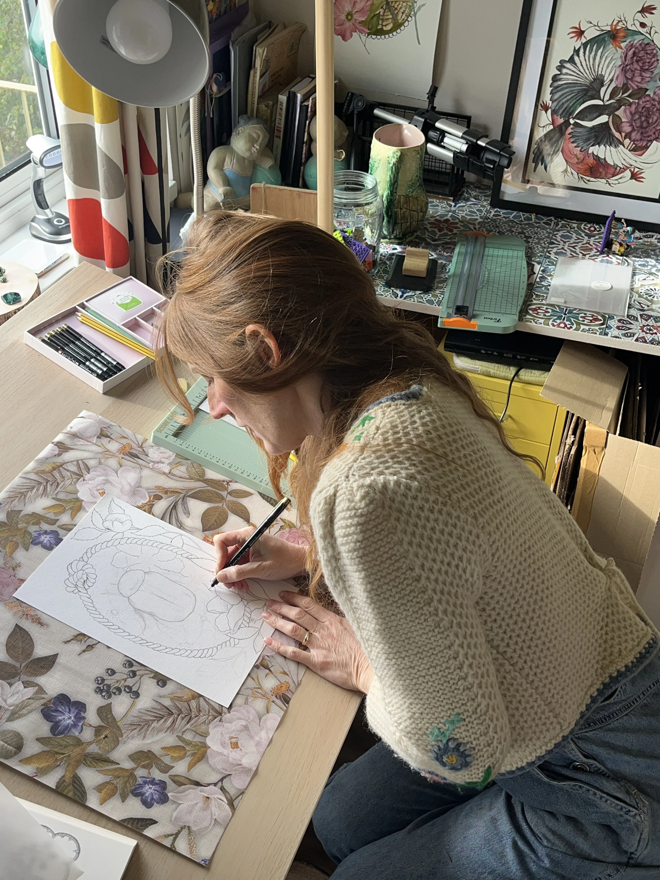 A woman is coloring a design on paper with floral and ornate elements, sitting at a worktable in a room filled with art supplies and decorations.