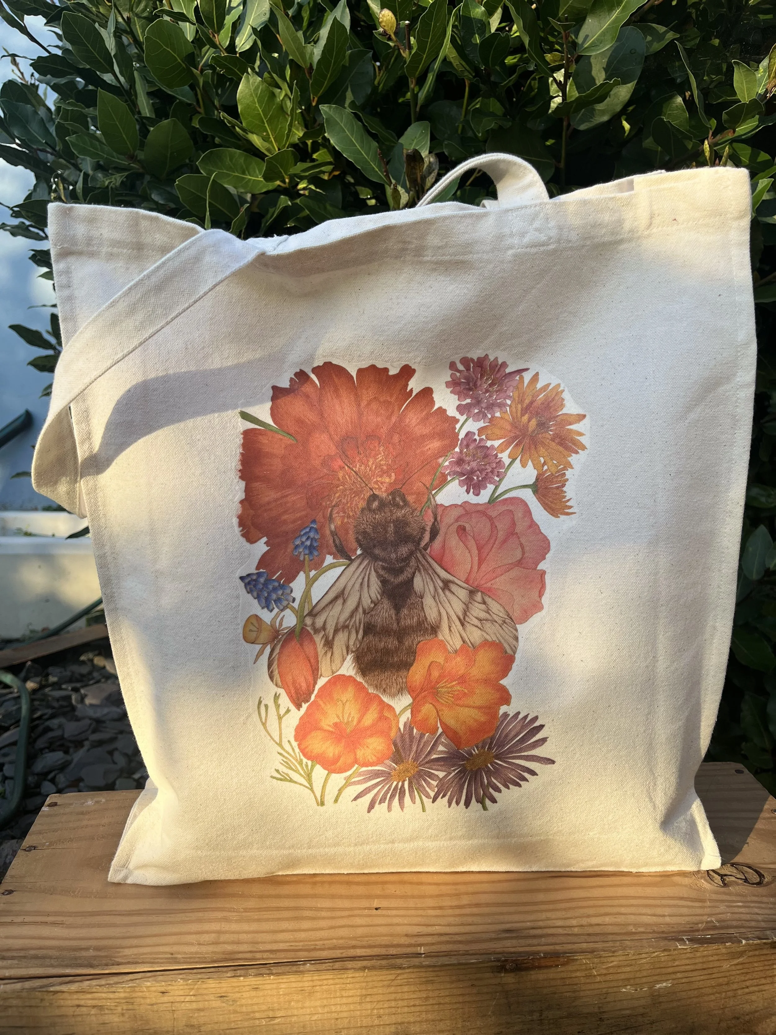 A cotton tote bag photographed outside with dappled sunlight. The design is of a bumblebee with bright orange and purple florals.