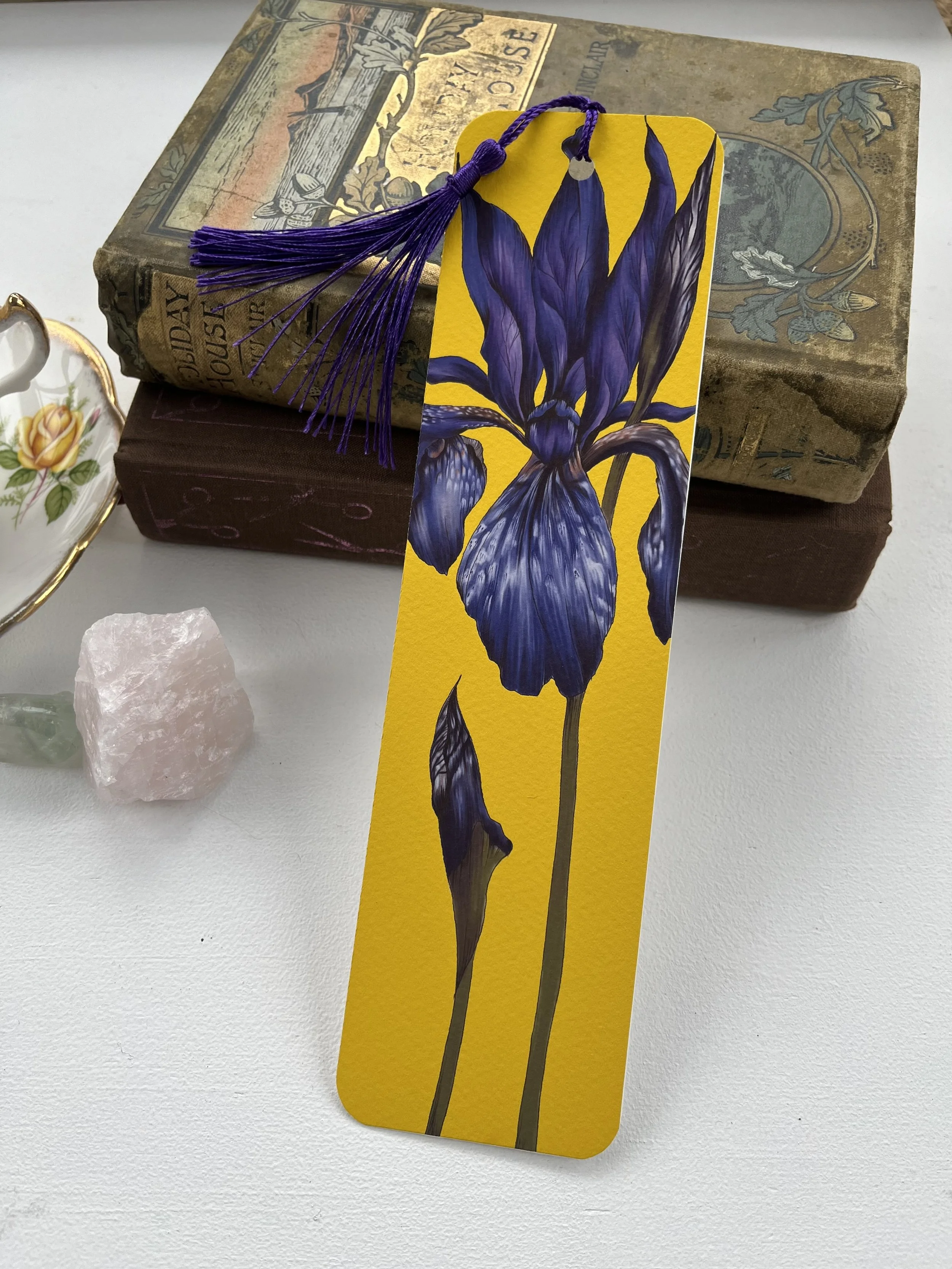 A yellow bookmark with purple iris flower leans against a stack of books. A teacup is visible in the background.