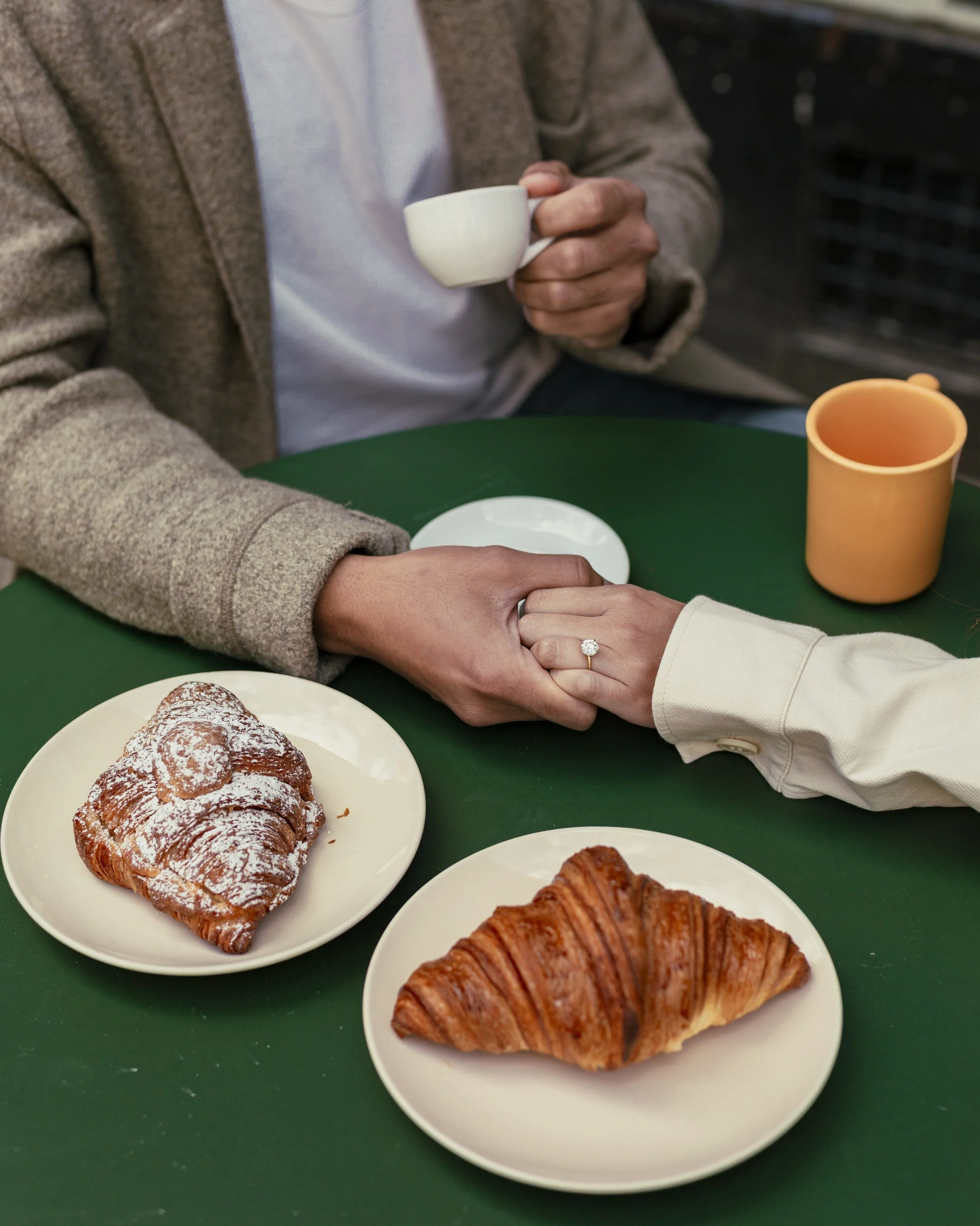Close detail of bride and groom holding hands at a café table with pastries and coffee.