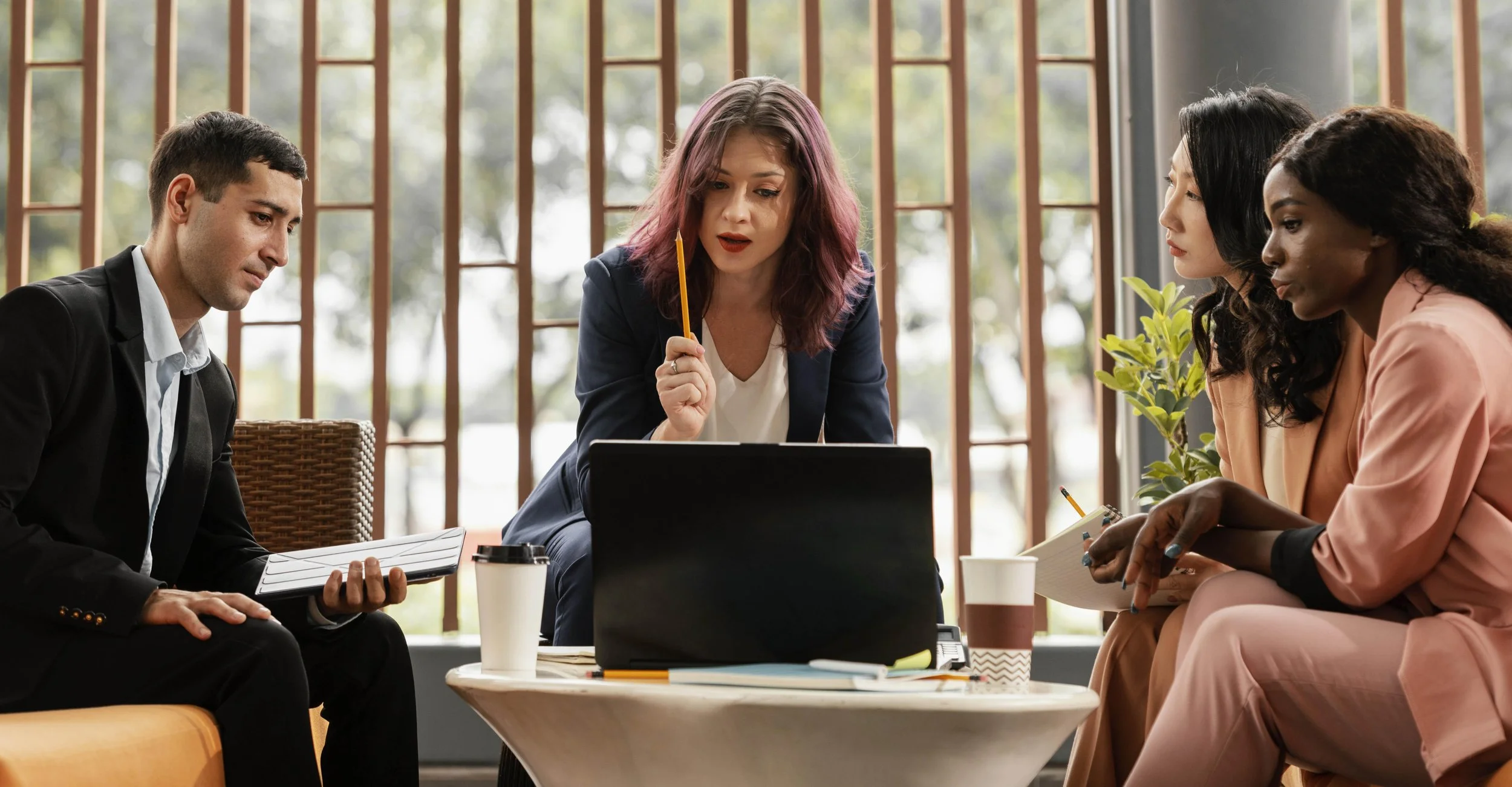 Four professionals in a meeting room having a discussion, sitting around a table with laptops, notebooks, and coffee cups, with large windows and green foliage outside.