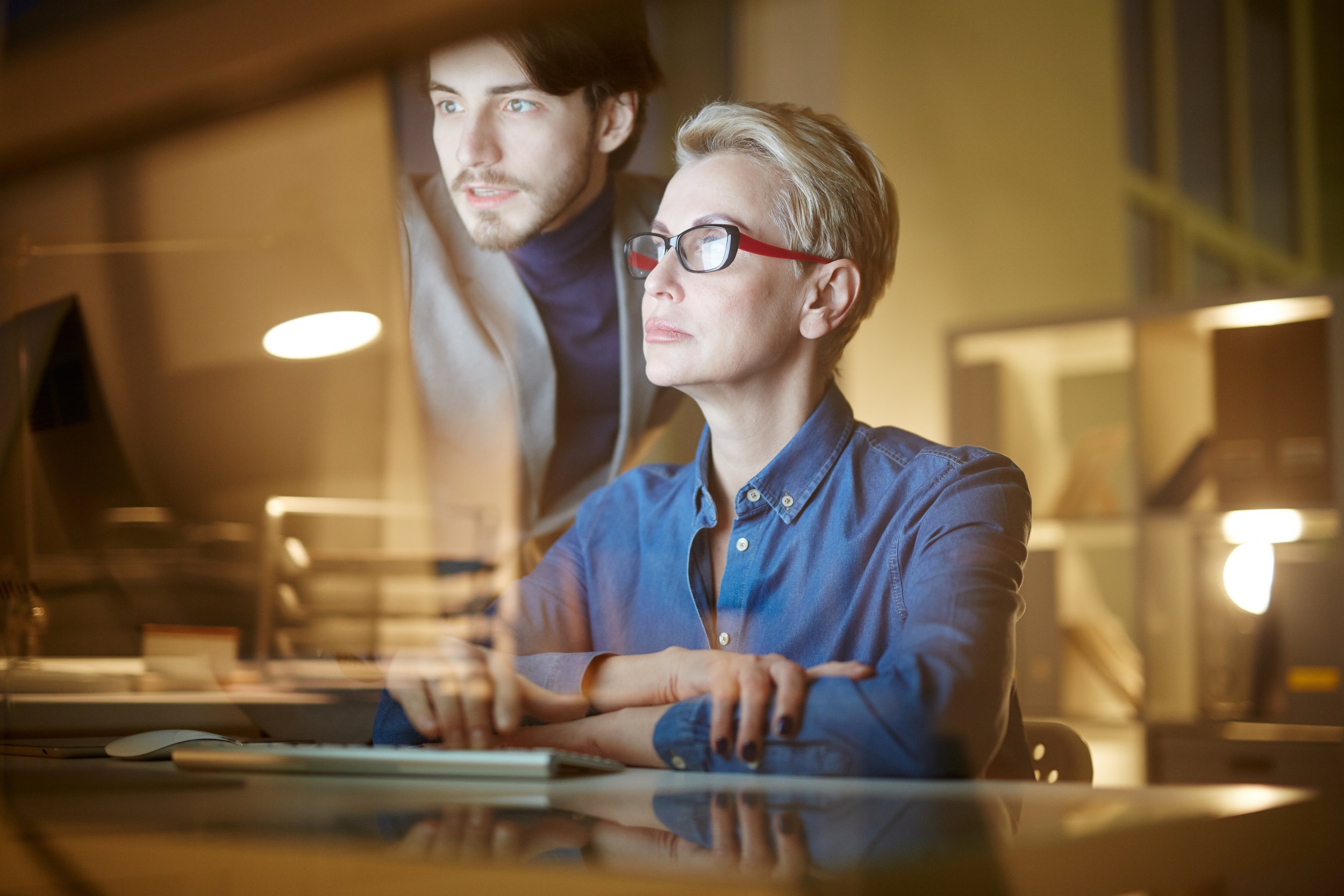 Two professionals working together in an office at night, looking at a computer screen with one sitting and the other standing behind, both focused.