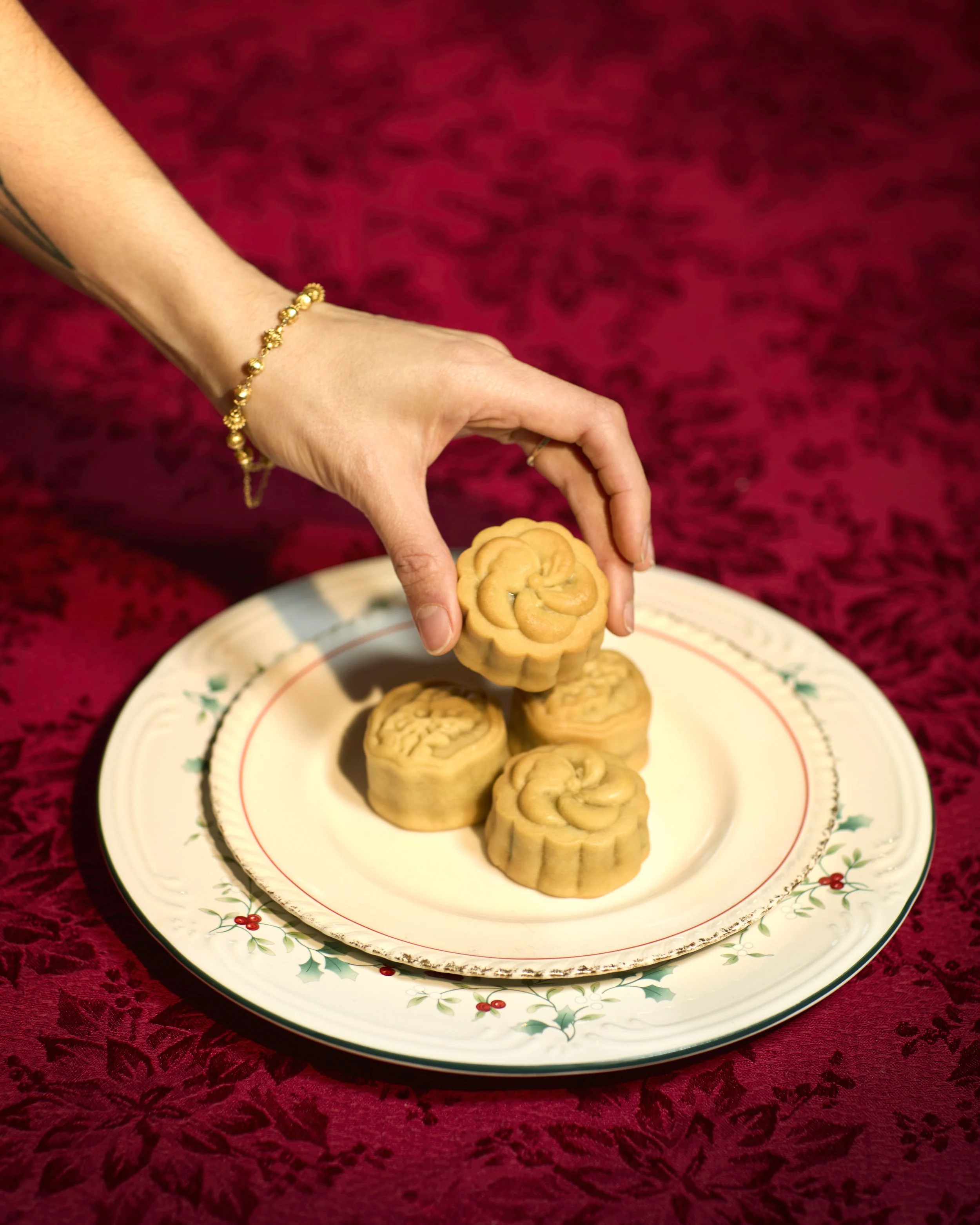 A hand wearing a gold bracelet picking up a mooncake with a floral pattern on a Christmas plate. Four more mooncakes are on the plate. The tablecloth is red with a velvet pattern.