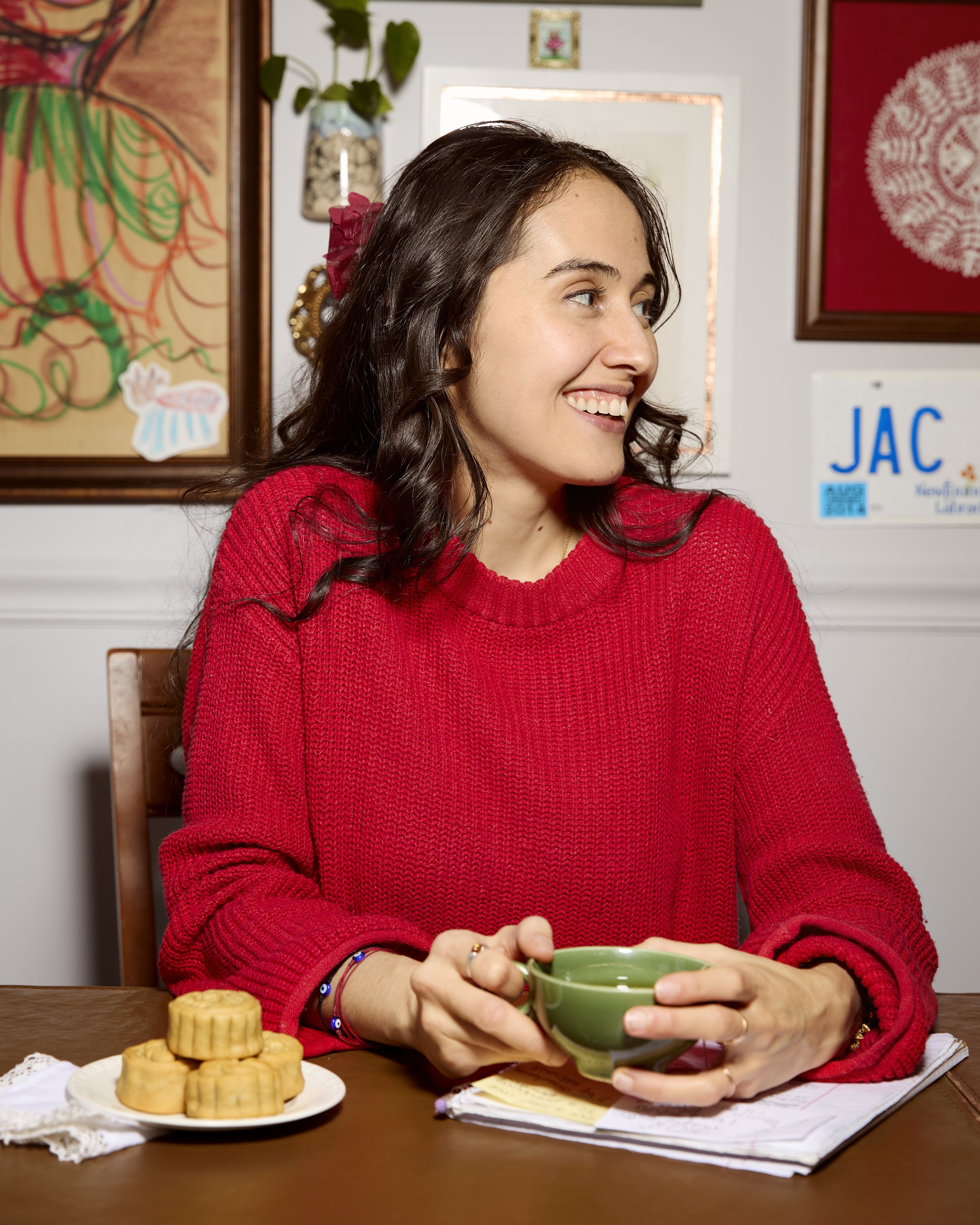 A young woman in a red sweater sitting at a table, smiling and holding a green mug. There is a plate of cookies and a notebook on the table. The background has colorful artwork, houseplants, and a sign with the word 'JAC'.