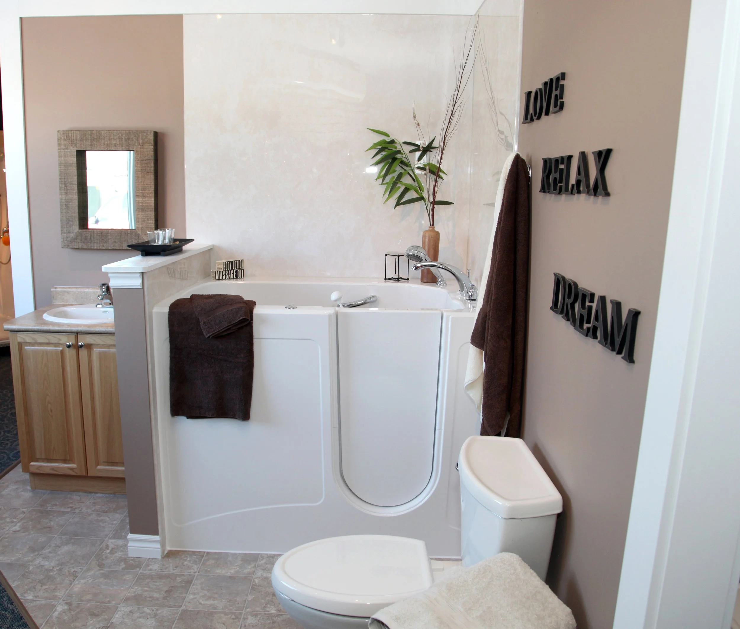 A bathroom with a walk-in bathtub, a toilet, a small sink with a wood cabinet, a mirror, decorative wall art with words "LOVE, RELAX, DREAM," a tall plant, and various towels.