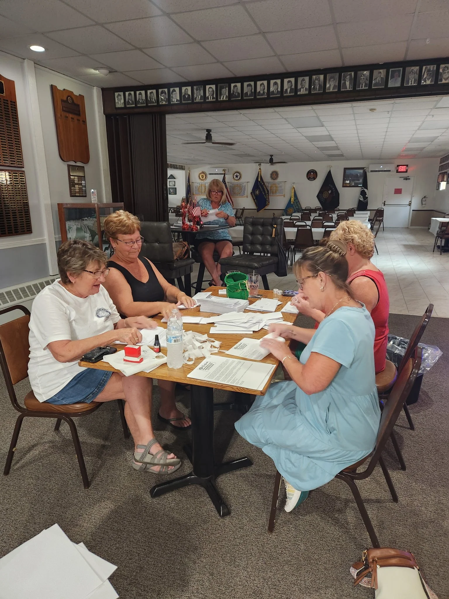 Five women sitting around a rectangular table in a room decorated with flags and framed photographs, reviewing papers and working on documents.