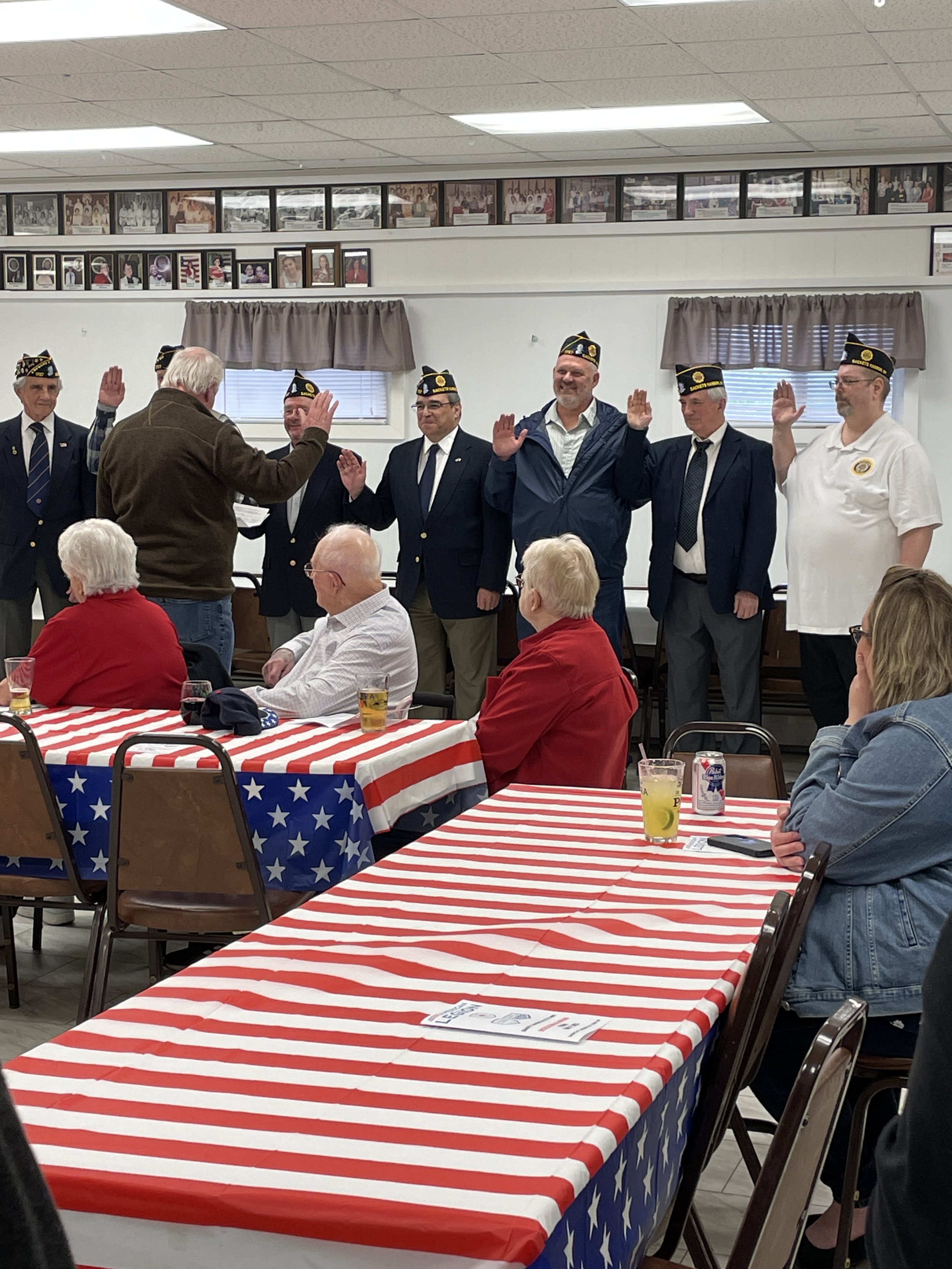 A group of men, some in military uniforms with hats, standing in front of an audience and raising their right hands during a ceremony or pledge, with tables covered in American flag-themed tablecloths.