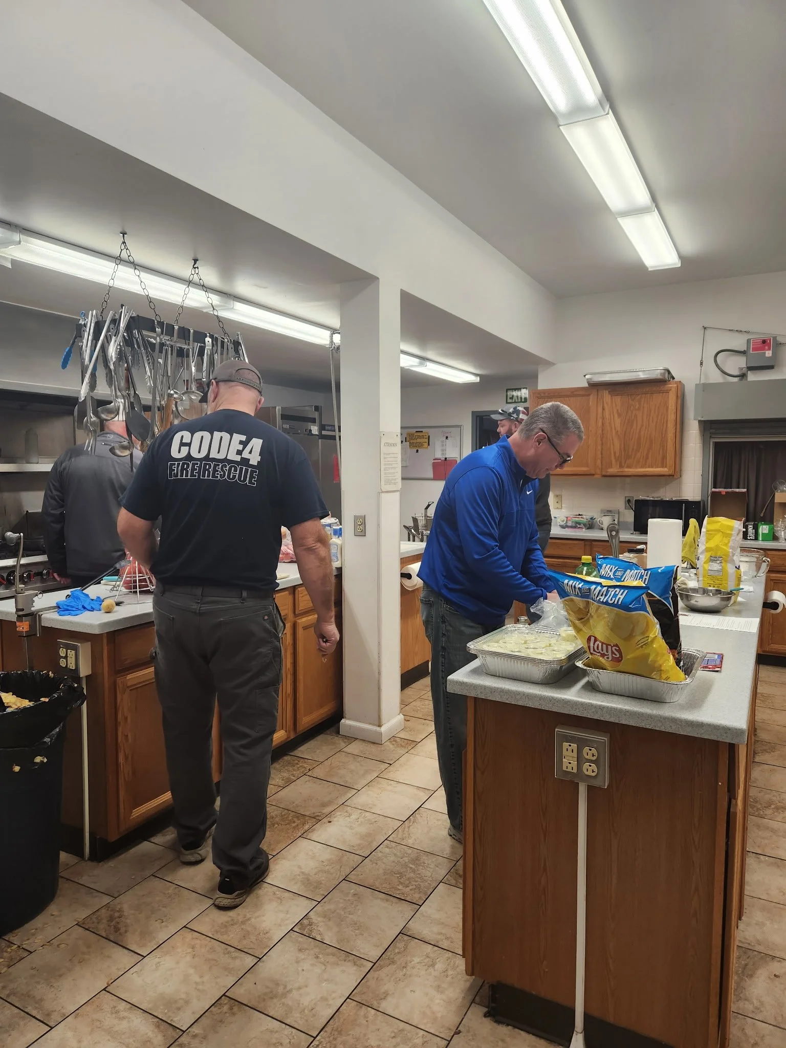 People preparing food in a kitchen, one wearing a 'CODE4 FIRE RESCUE' t-shirt, and another mixing ingredients on the counter, with cooking tools hanging above.