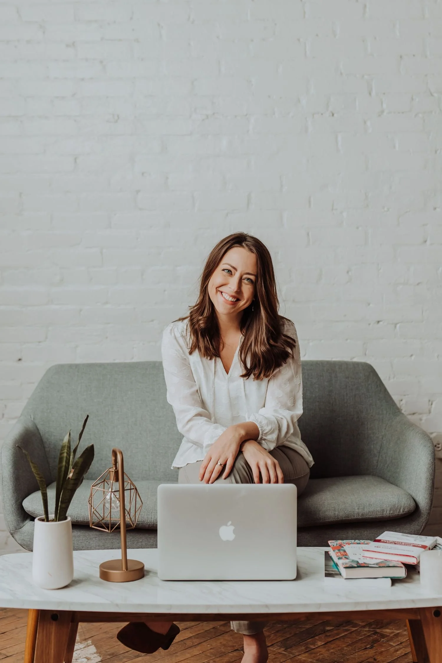 A smiling woman with brown hair sitting on a gray sofa behind a white coffee table, with a laptop, books, a potted plant, and a geometric copper desk lamp in a bright room with a white brick wall.