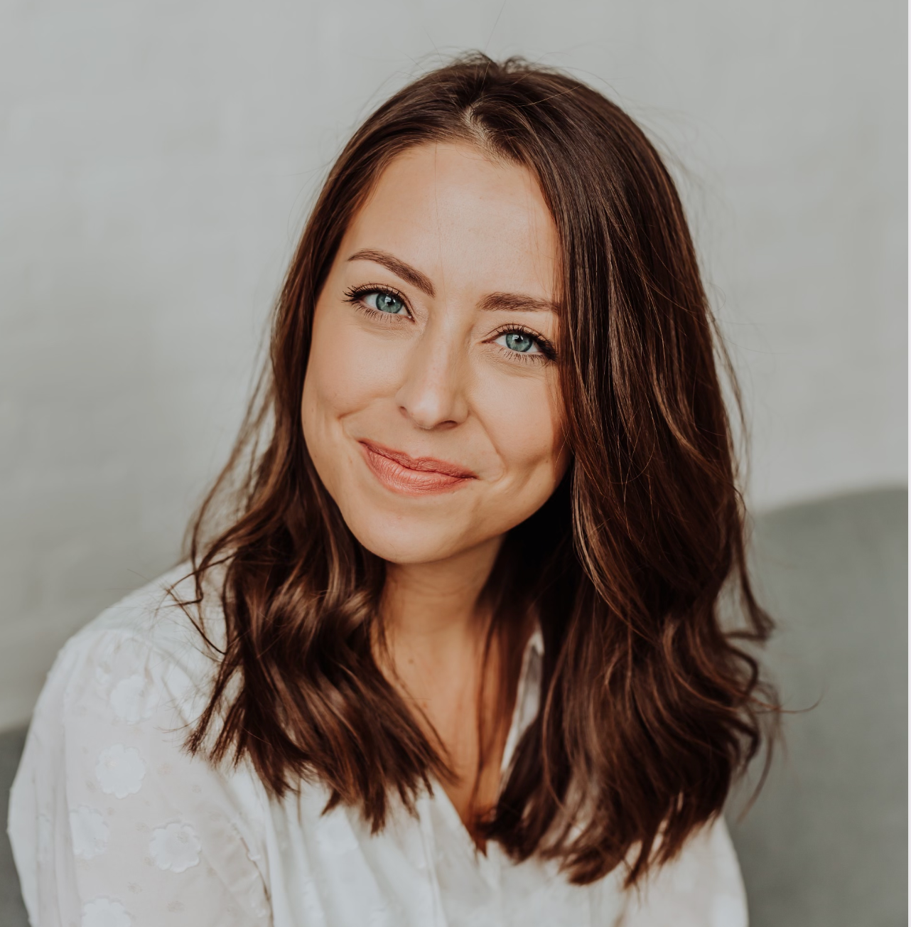 Close-up of a woman with long wavy brown hair, blue eyes, and a friendly smile, sitting against a light-colored wall.