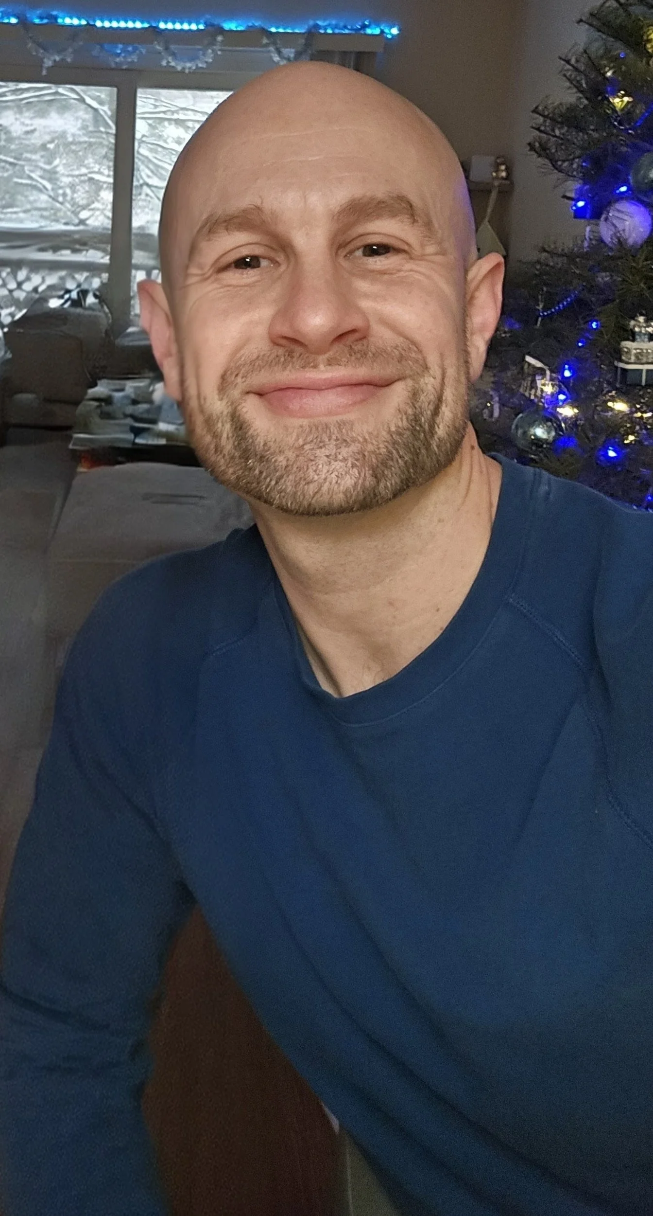 A smiling man with a beard and no hair, wearing a dark blue long-sleeve shirt, sitting indoors in front of a decorated Christmas tree with blue lights and ornaments.