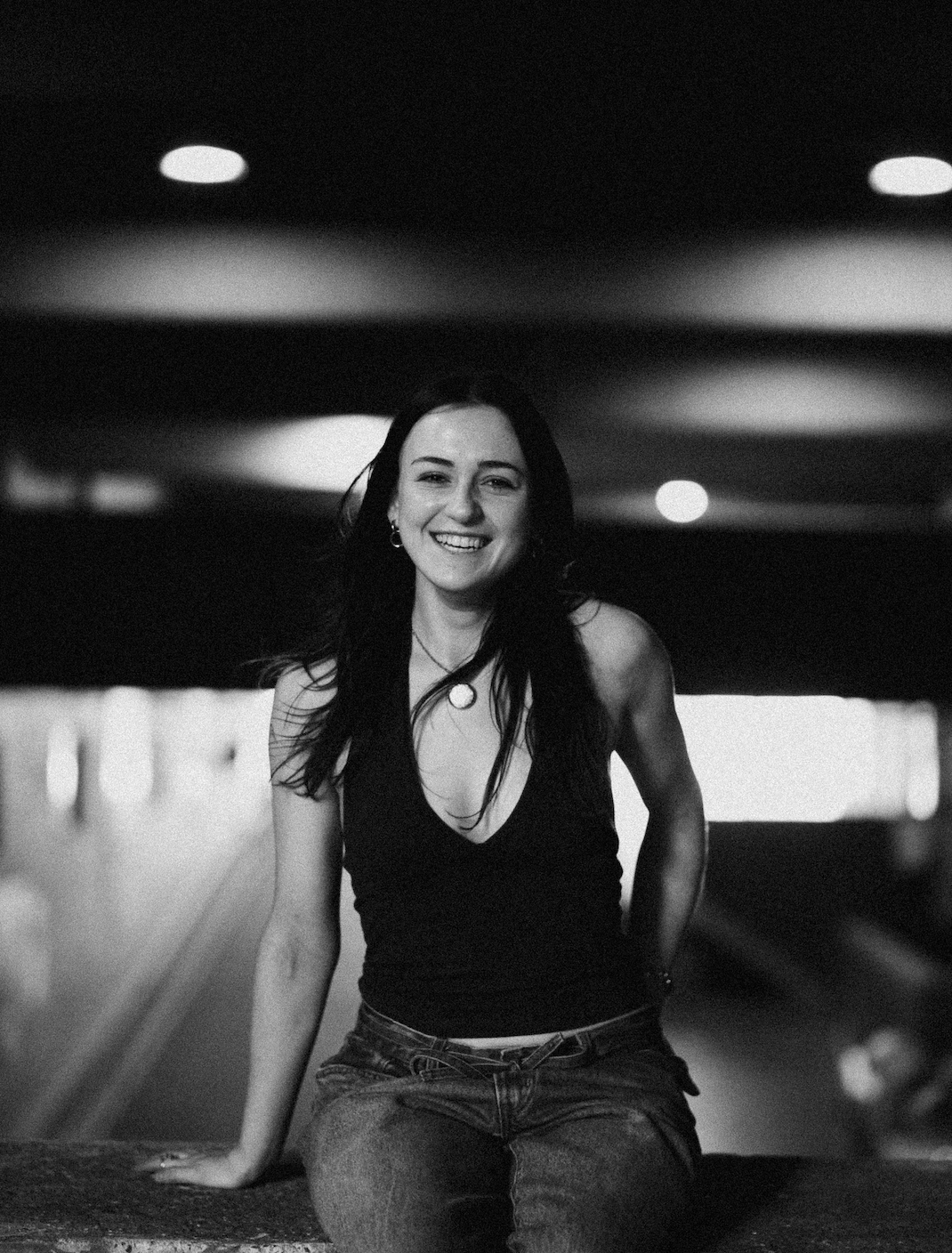 Black and white photo of a young woman with long dark hair, smiling, sitting on a ledge in an underground parking garage.