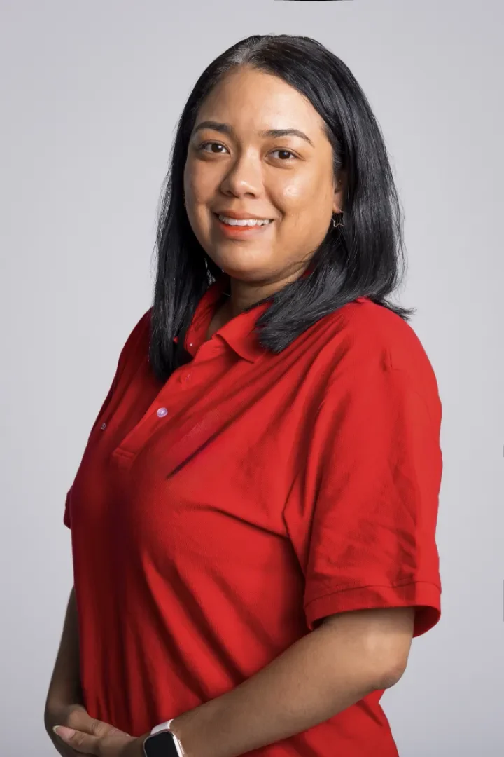 A woman with black hair wearing a red polo shirt, smiling, standing against a plain gray background.