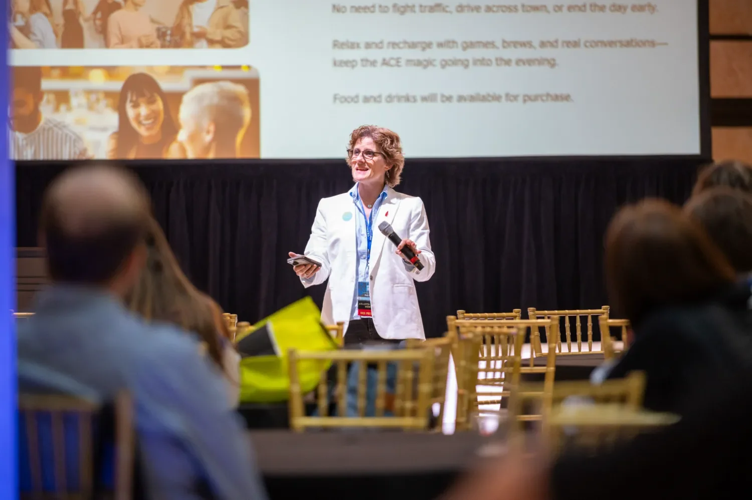A woman in a white blazer speaking at a conference, holding a microphone and a remote, in front of a large projection screen with text and images, audience members seated at tables in the foreground.