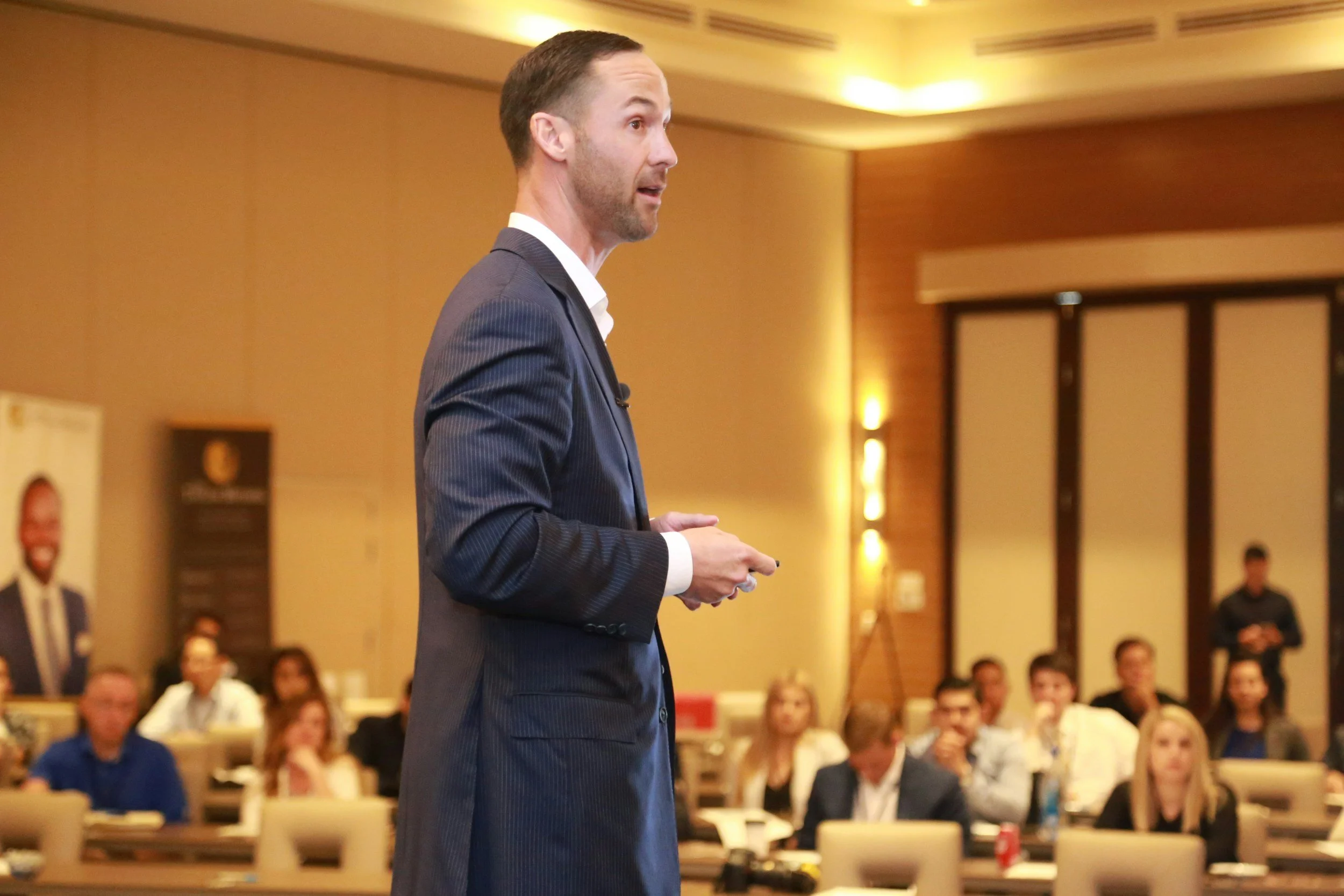 A man in a dark suit giving a presentation to an audience in a conference room with warm lighting.